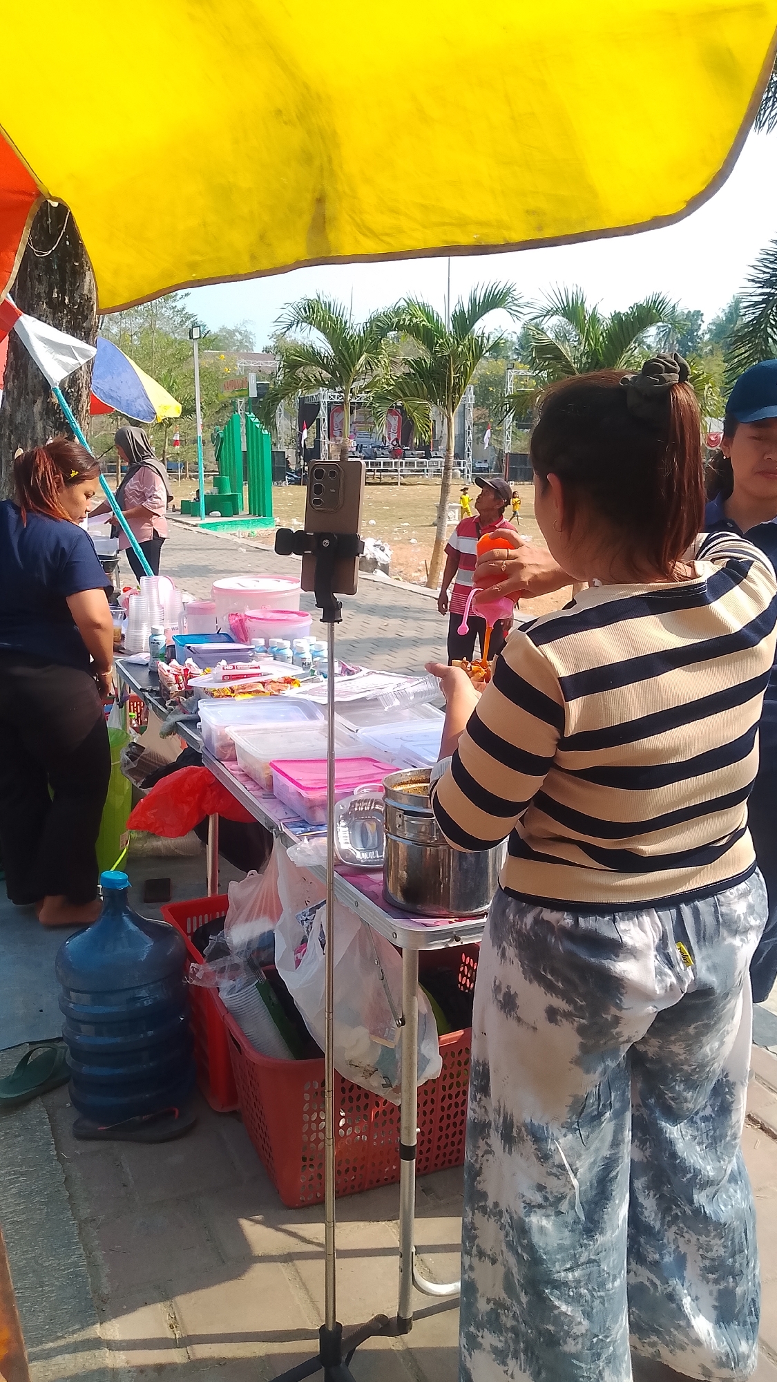 An outdoor scene in Karanglangu, Indonesia, shows a stall with food items and drinks. A woman in a striped shirt and patterned pants is preparing food, holding a bottle and a plate. Another woman in a dark shirt and dark pants is standing behind the stall, looking down. In the background, a stage with a band is set up, and palm trees are visible. Several people are present, some standing and some sitting. A large blue water jug is placed on the ground next to the stall. The scene appears to be part of a local event or festival.