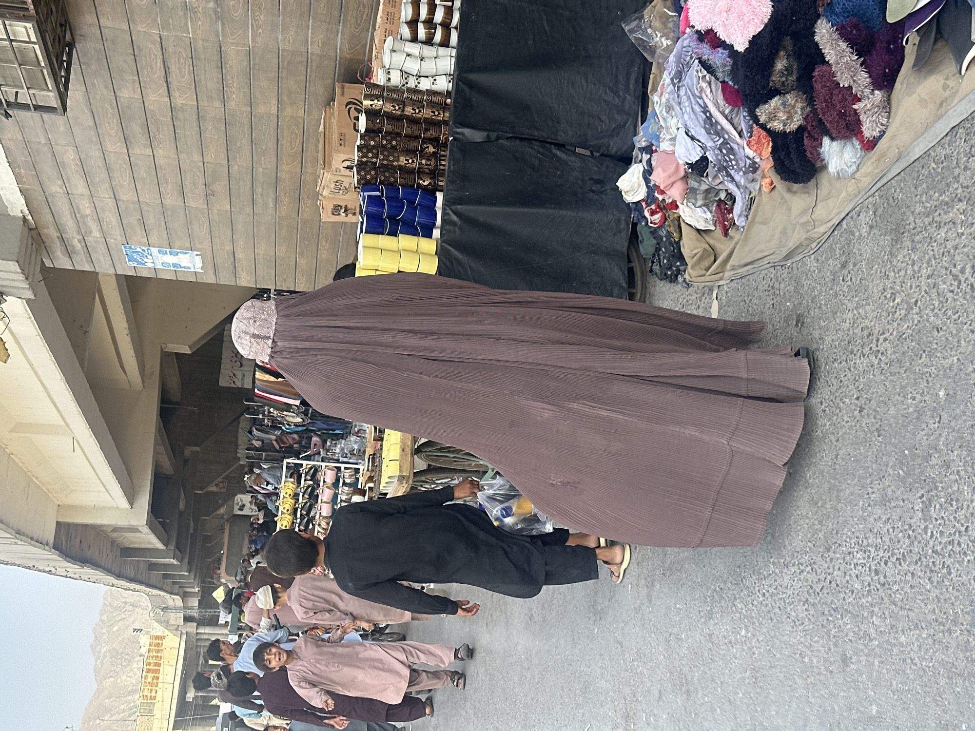 The image captures an outdoor market scene in Quetta City Tehsil, Pakistan. A woman wearing a full-body brown burqa is walking past a market stall. The stall displays items for sale, including cups in boxes and various textiles. Several males, mostly dressed in traditional clothing, are also visible. The background features buildings and a bridge structure, while mountains are visible in the far distance. The overall atmosphere is one of a bustling marketplace.