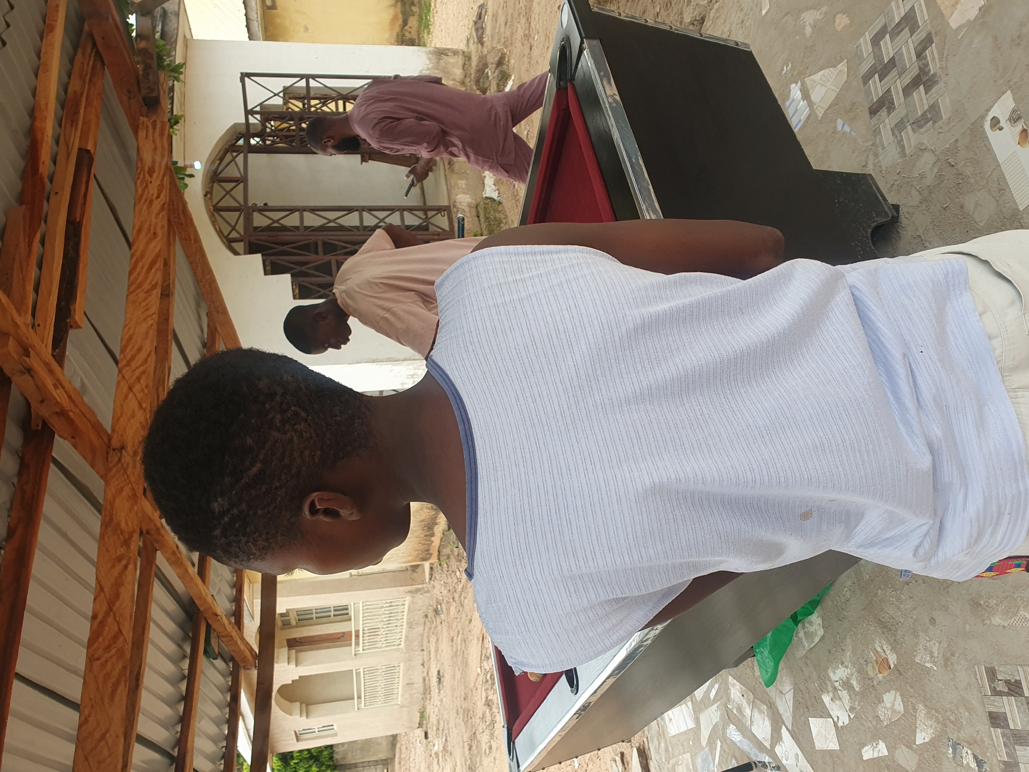 In Lokoja, Nigeria, three individuals are present outdoors. In the foreground, a person wearing a light blue striped sleeveless shirt stands with their back to the camera, facing a pool table. To the left of the pool table, a red pool ball is visible. In the middle ground, a person in a light tan t-shirt is seen from the side, facing the pool table. Further in the background, a person in a maroon traditional Nigerian outfit is walking away, holding a mobile phone. They are walking towards a white building with an arched entrance and metal gate. The environment appears to be an unpaved area with scattered debris and some decorative tiles on the ground. A wooden-framed roof with corrugated metal sheeting covers the area.
