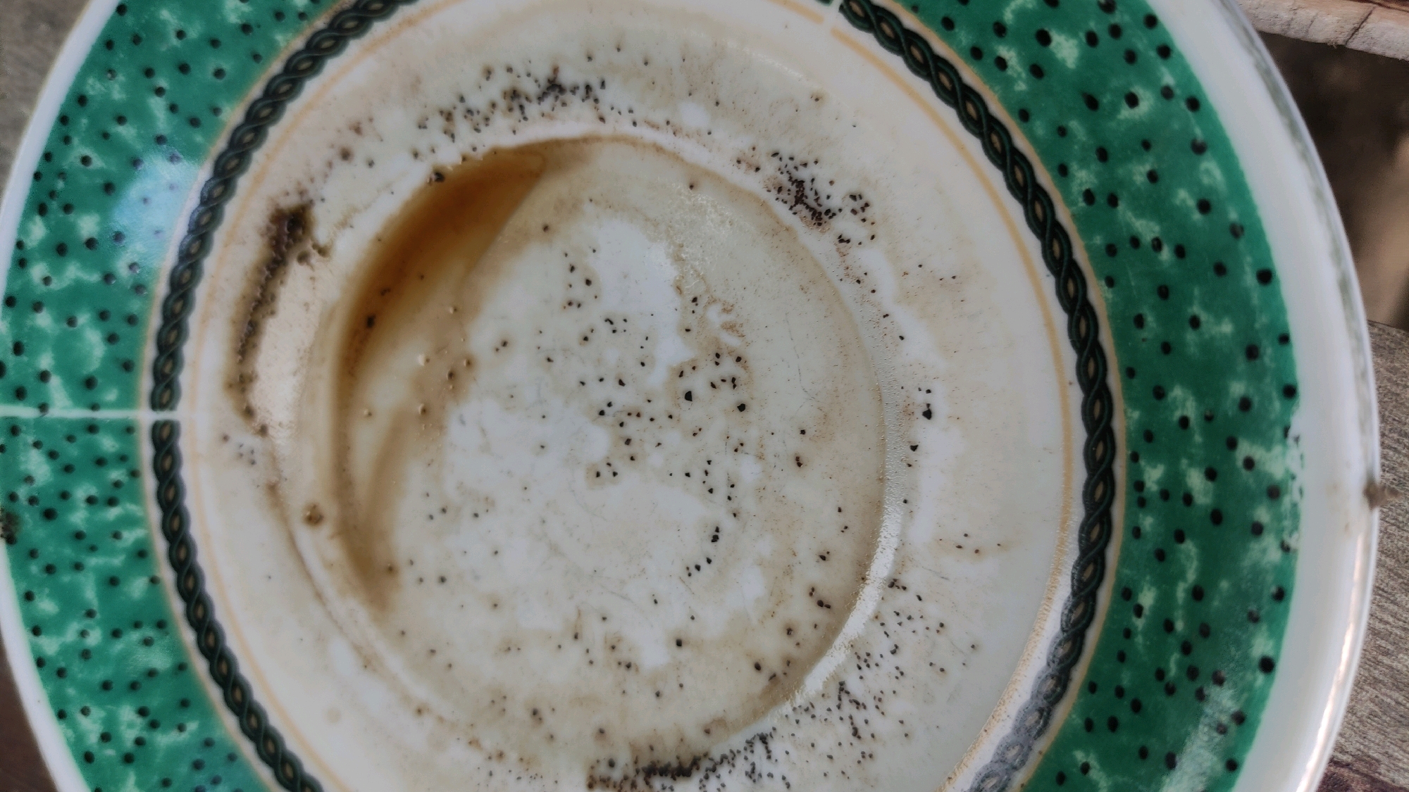An overhead, close-up, slightly angled shot shows the bottom of a dirty white ceramic plate. The plate has a decorative rim featuring a braided black and white pattern, followed by a wide band of green with evenly spaced black dots. In the center of the plate, there are remnants of a brown liquid, likely coffee, with visible coffee grounds scattered throughout. The plate is resting on a wooden surface. No people or animals are visible.
