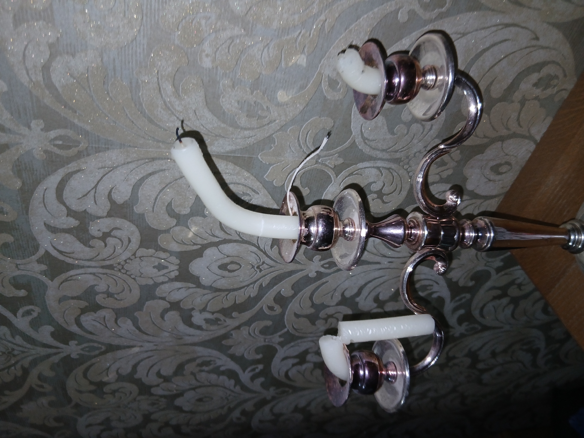 A close-up, eye-level shot shows a metallic candelabra with three candles, two of which are broken. The candelabra is positioned on a wooden surface in front of a patterned wallpaper. The wallpaper has a damask design in shades of light grey and beige with some subtle glitter. The candelabra is silver or chrome and has ornate detailing. The candle on the left arm of the candelabra is broken into two pieces, with the top piece detached. The candle on the right arm is also broken, with the top portion bent over. The candle on the center arm is intact but appears to be unlit. The lighting in the scene is dim, casting shadows and highlighting the metallic sheen of the candelabra and the glitter on the wallpaper. There are no people present in the image. The location of the scene is identified as Shymkent, Kazakhstan.
