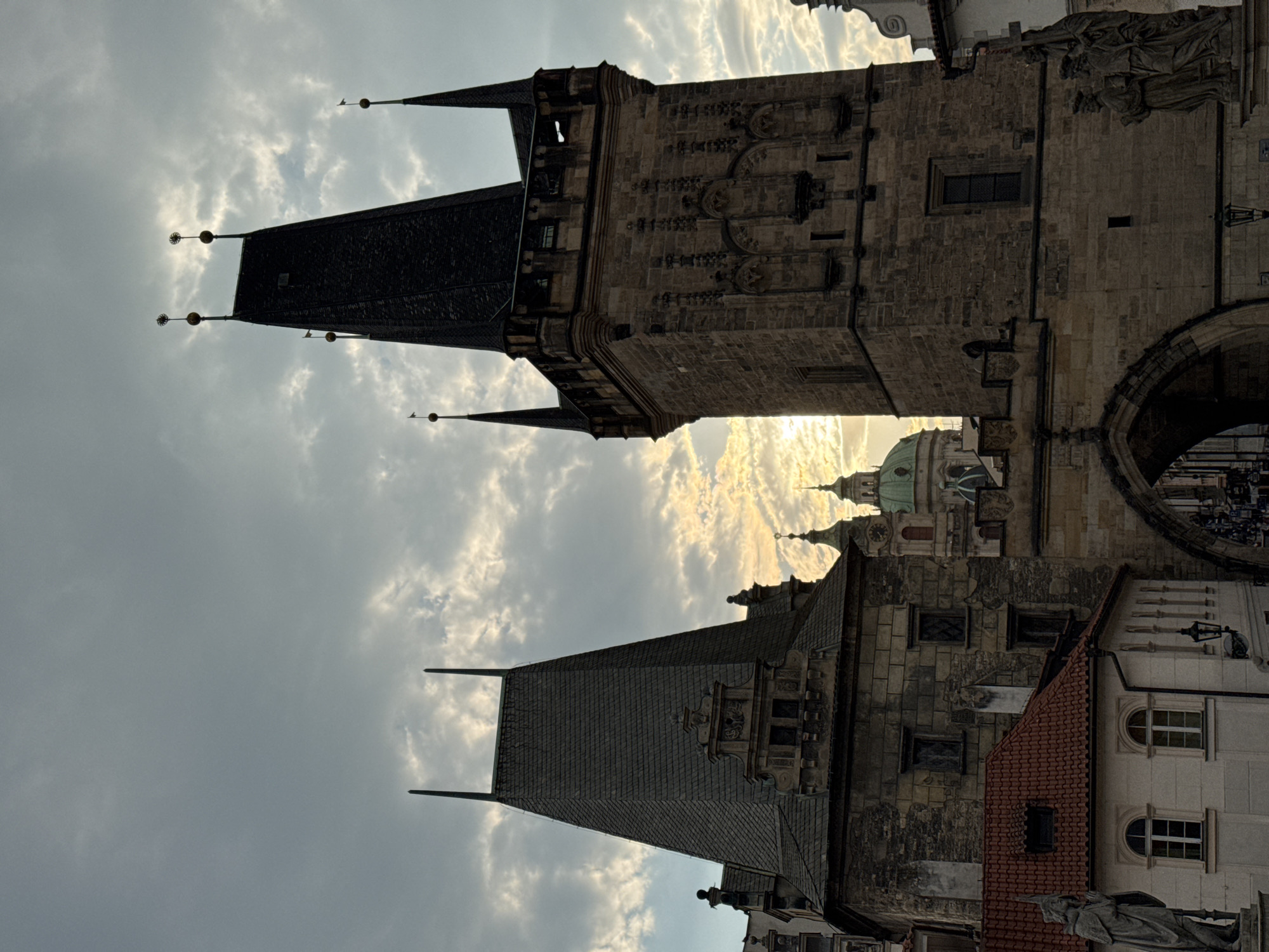 This image captures a view of the Old Town Bridge Tower and the Lesser Town Bridge Tower in Prague, Czechia, at dusk. The sky is filled with dramatic clouds illuminated by the setting sun, creating a warm glow behind the gothic architecture. In the foreground, the stone structures of the bridge towers rise majestically, their dark, steep roofs contrasting with the lighter stone walls. Intricate carvings and architectural details are visible on the towers. To the left, a smaller building with a red-tiled roof and arched windows is visible, with a statue of a robed figure standing to its left. In the background, the domes of other historic buildings, possibly churches, are visible. The overall scene evokes a sense of historical grandeur and the beauty of Prague at twilight.