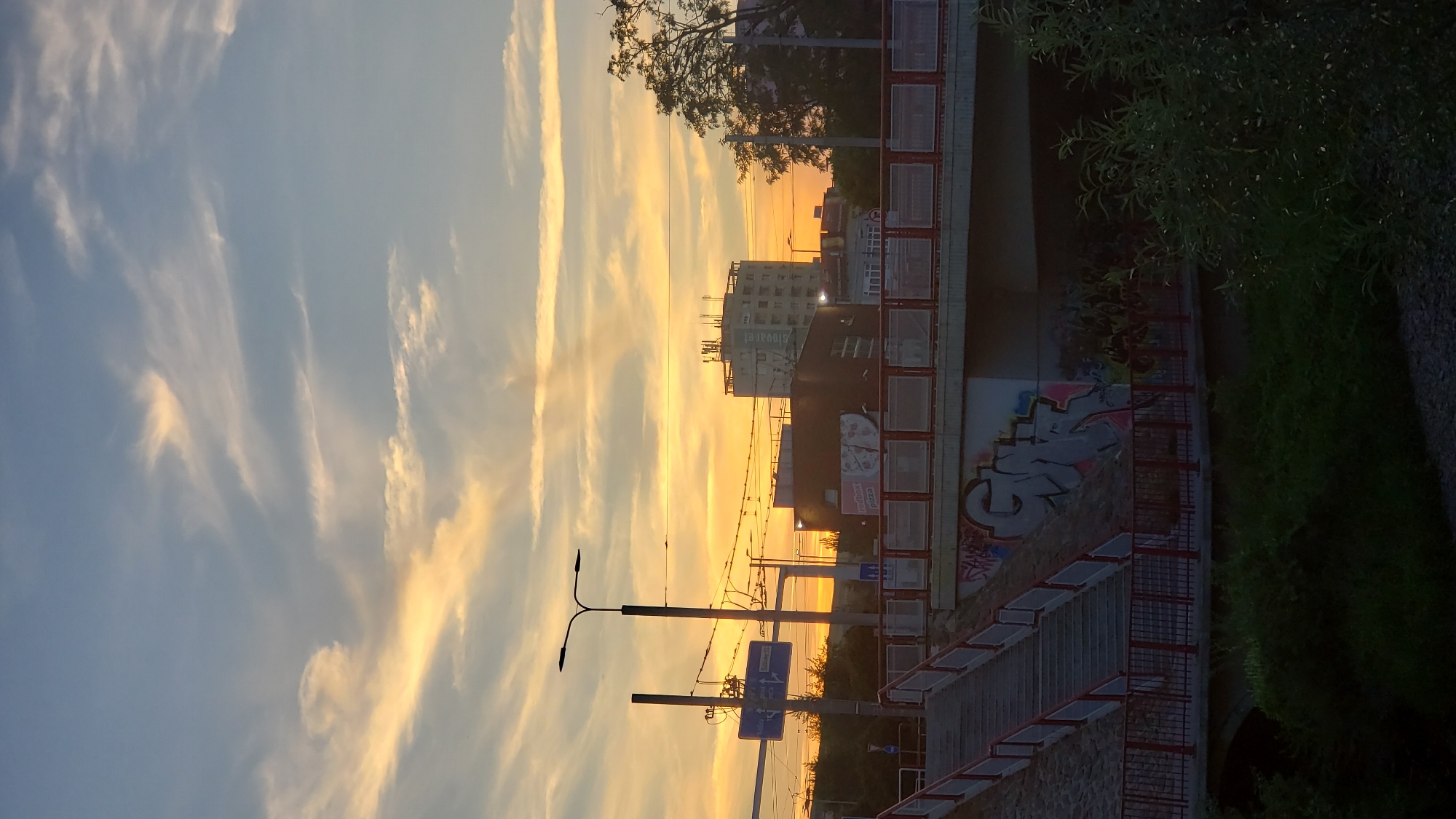 The image captures a sunset over a city, likely Prešov, Slovakia, indicated by the street signs. In the foreground, a set of stairs with red railings leads up to a bridge. The bridge itself is adorned with graffiti on its underside, visible behind a red fence. Beyond the bridge, a street scene unfolds with traffic lights, power lines, and several buildings. One prominent multi-story building in the background features a sign with the name "Slavart". The sky is a canvas of warm sunset colors, with streaks of orange, yellow, and pink clouds against a soft blue. Lush green foliage frames the bottom of the image, suggesting a natural element within the urban landscape.