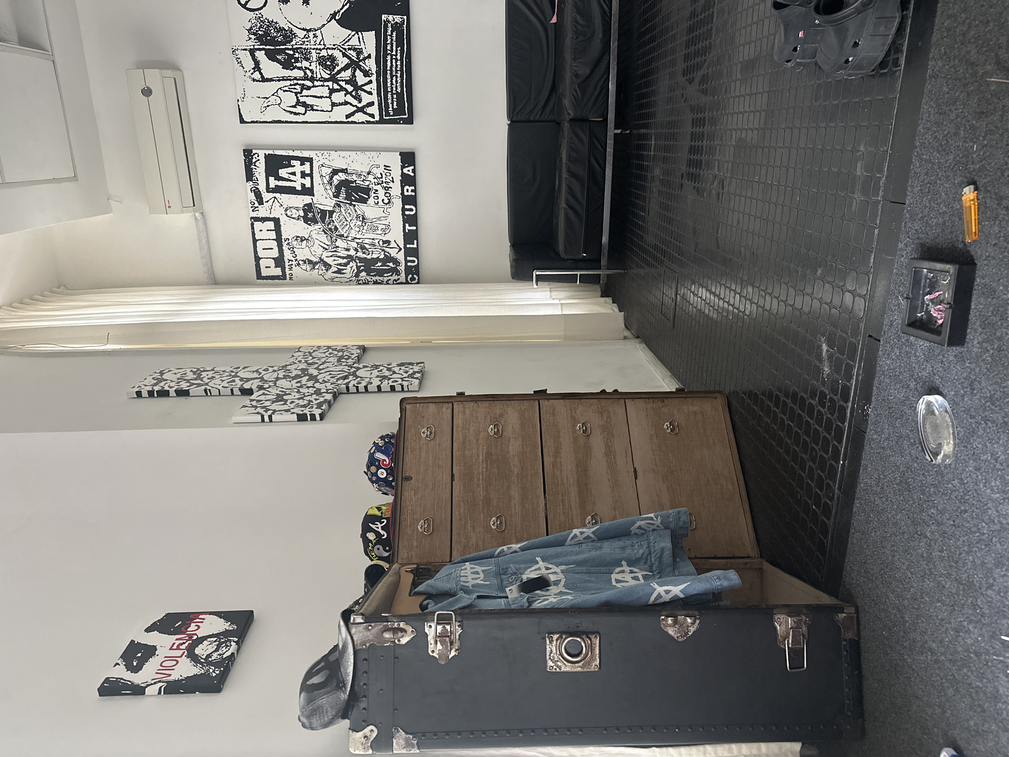 The scene appears to be an interior shot, potentially a living space or studio, featuring an eclectic mix of items and decor. The wall is white, adorned with black and white graphic art. A large trunk-like chest sits on the floor, partially open, with a denim jacket inside. An ash tray with a lighter sits on the floor. The flooring is dark and patterned, possibly tile or linoleum. The photograph seems to be taken from an elevated perspective, focusing on the juxtaposition of the artwork and the chest's contents. The image has a modern, artistic vibe. The location in Buenos Aires, Argentina is not determinable from this image.