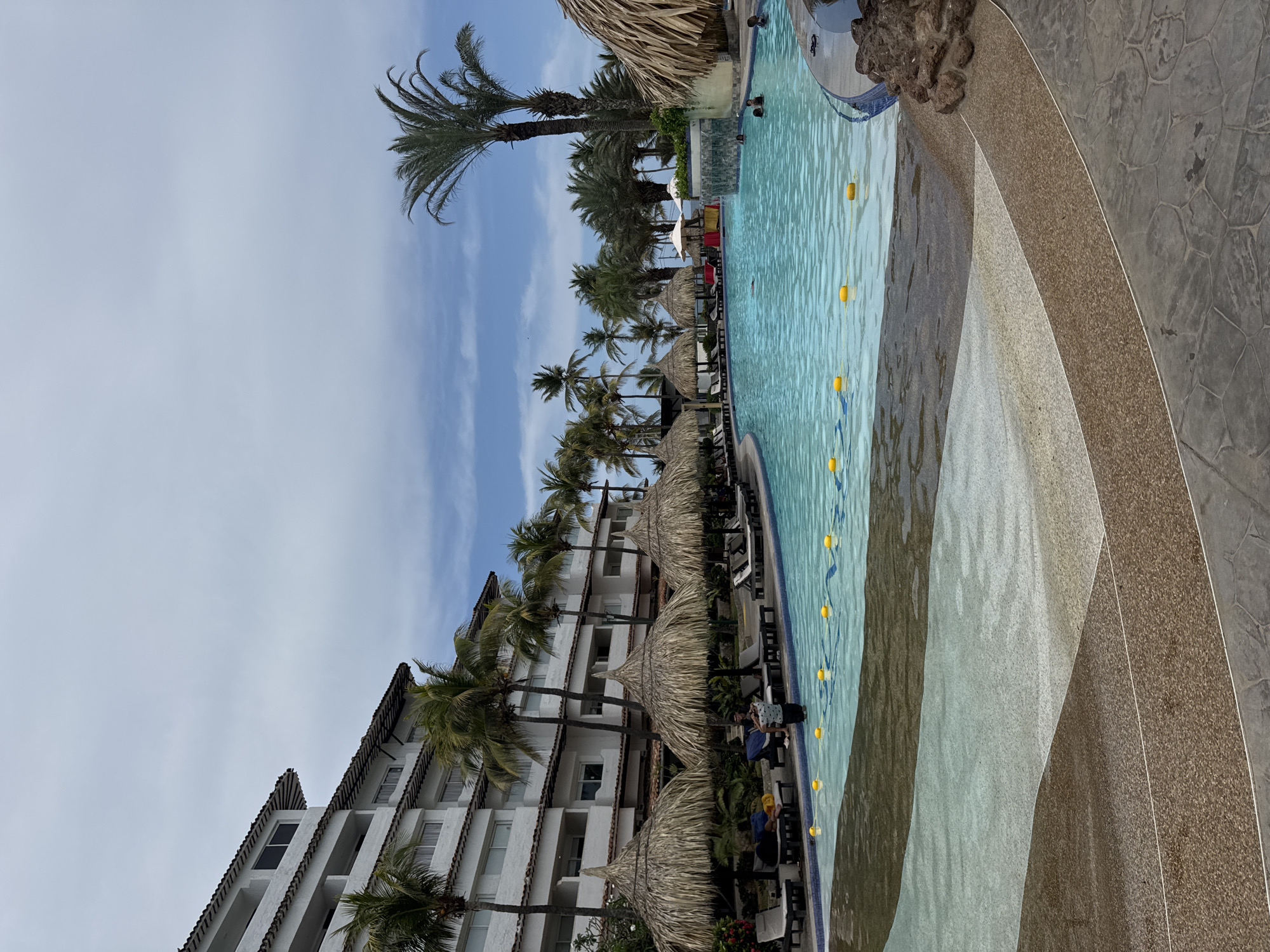 The image depicts a resort pool in El Tirano, Venezuela. A multi-story white building with dark tiled roofing is lined with palm trees and thatched-roof umbrellas alongside the pool. Yellow buoys float in the pool. Lounge chairs are visible beneath the umbrellas. A few people are seen near the pool, and there are swimmers in the pool. The pool's edge has a tiered, textured stone design. The sky is partly cloudy.