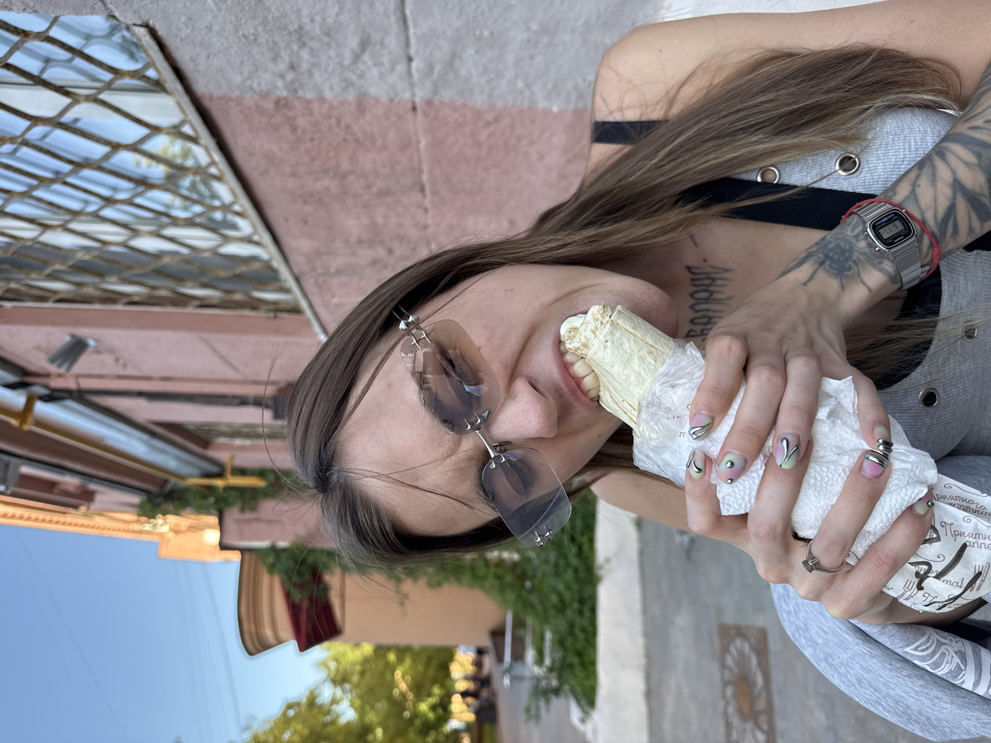The image captures a young female subject in an outdoor urban environment, specifically identified as the city of Moscow, Russia. The subject, positioned in the foreground and center, possesses long, straight, medium-brown hair and is wearing rimless sunglasses with small silver stud details on the temples. She is engaged in the act of consuming a large wrap, consistent with a shawarma, aggressively biting into it with her mouth wide open, exposing her teeth. The wrap is partially encased in a white paper wrapper featuring black Russian text, including the word "Приятного" (Enjoy).
Her attire consists of a black tank top layered beneath a light grey ribbed top accented with silver eyelets. Visible tattoos include a detailed dark insect or beetle-like design and botanical patterns on her right forearm, and black gothic script reading "Бананы" (Bananas) on her upper chest/collarbone area. On her left wrist, she wears a silver digital watch (Casio A158W-1) over a thin red string bracelet. Her left hand is adorned with several silver rings, including one with a dark square stone on her ring finger and a coiled design on her pinky finger. Her fingernails are almond-shaped and feature a gradient manicure transitioning from pink to light green, embellished with abstract silver metallic swirls and small silver beads.
The background consists of a multi-story building with a light reddish-pink facade, likely stucco or painted brick. Yellow utility pipes are visible along its exterior. An older window with a distinct decorative metal grate is present on the building's right side, reflecting the clear blue sky and blurred green foliage. Towards the upper left, an orange-brown cornice and a reddish-brown awning are visible. The upper left portion of the image shows a clear blue sky. In the distant left background, blurred green trees and indistinct human figures suggest a park or pedestrian zone. The ground surface is a light-colored paved material, indicative of a sidewalk or public square.