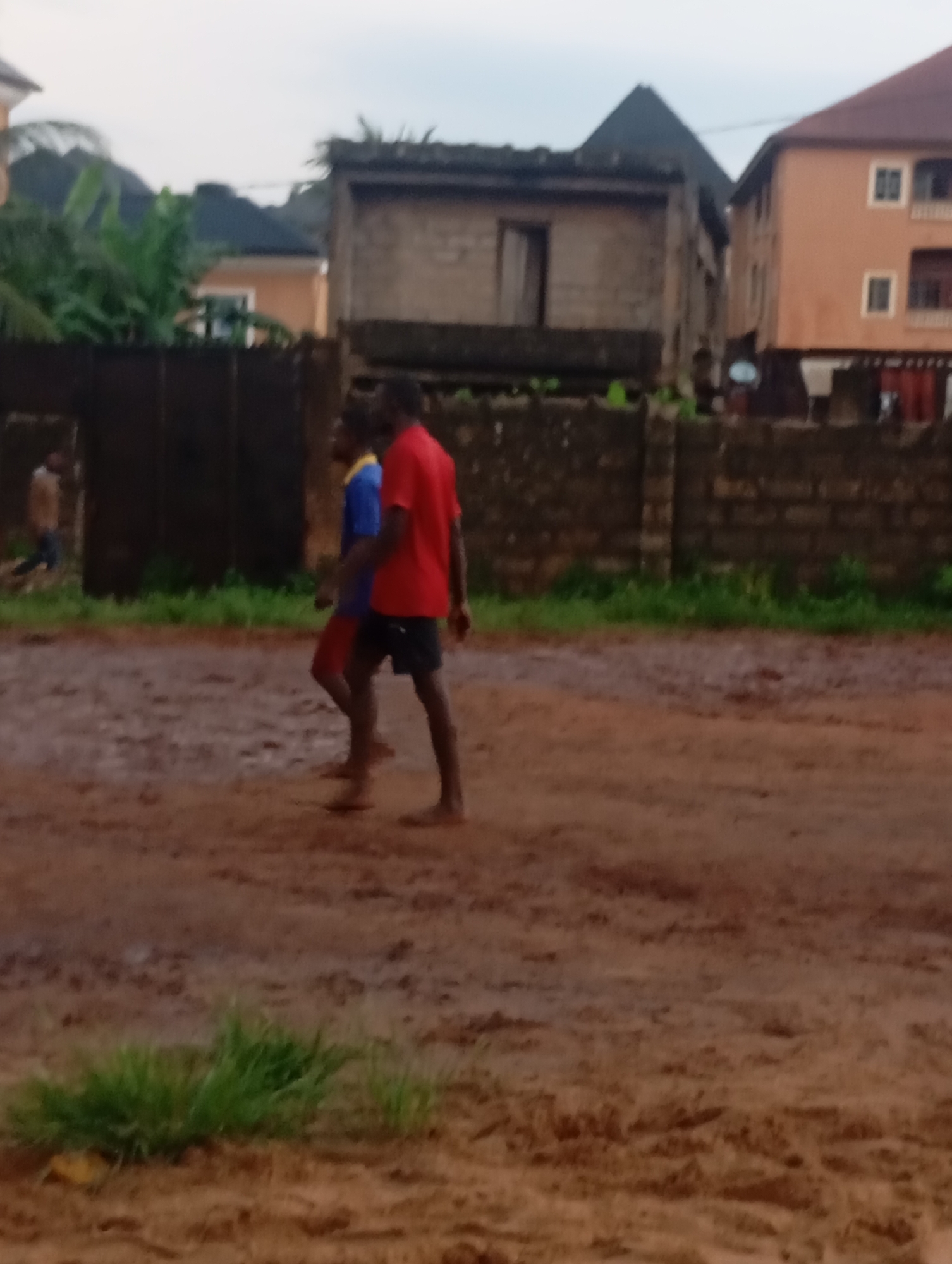 Two barefoot individuals, one wearing a red shirt and the other a blue shirt, walk side-by-side on a muddy surface in Akpaka Forest Reserve, Nigeria. A third individual walks away from the viewer near a metal fence. Behind the fence are brick buildings, some with corrugated roofing. Some green vegetation is visible.