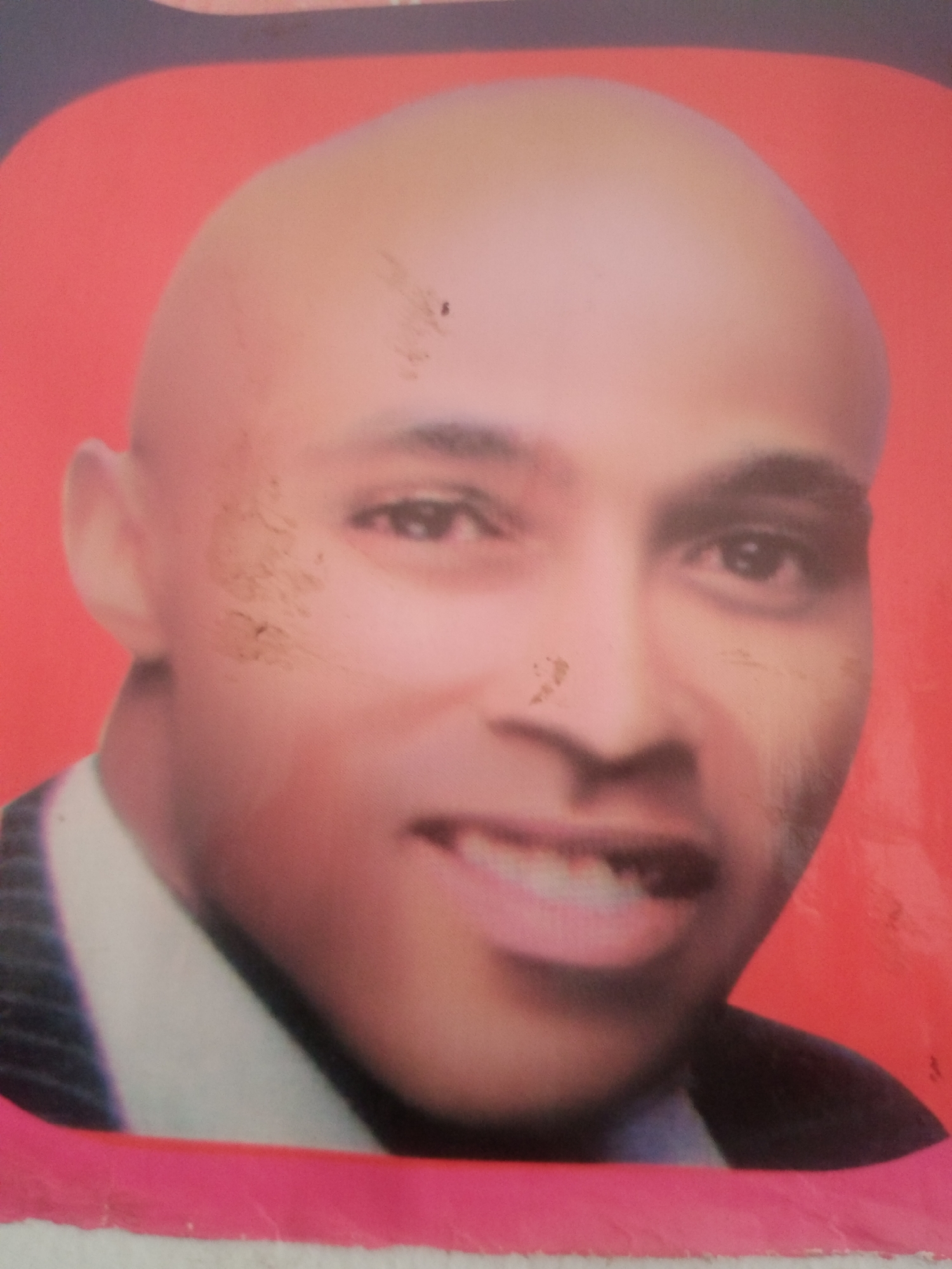 A portrait photograph of a bald, light-skinned man with dark eyes and eyebrows, wearing a white collared shirt and a dark suit jacket. He has a slight smile, revealing his teeth. The background is a solid reddish-orange. The photograph appears to be old or has been damaged, with several small specks and smudges visible on the surface. The man's face is centrally framed. This image does not contain any recognizable geographical landmarks or indicators that would allow for its precise location within Jalingo, Nigeria.