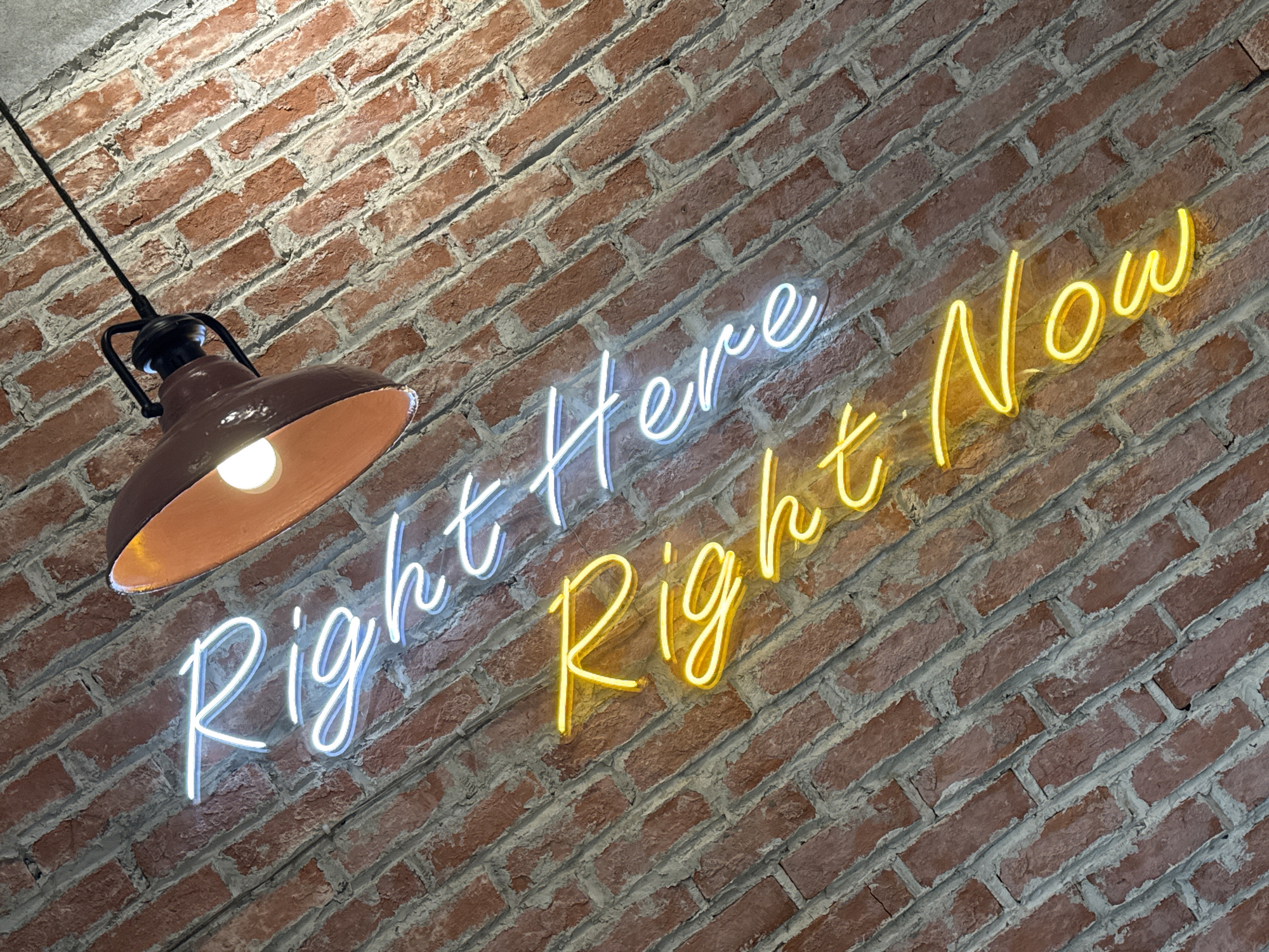 This is a close-up, angled shot of a brick wall in Da Nang, Vietnam. Two lines of neon text are mounted on the wall. The top line, in white neon, reads "Right Here". The bottom line, in yellow neon, reads "Right Now". A single light fixture with a copper-colored shade hangs from the upper left of the frame, casting a warm glow on the brick and illuminating the neon signs. The bricks are a reddish-brown with visible mortar lines, and the overall lighting suggests an indoor setting, possibly a cafe or bar.