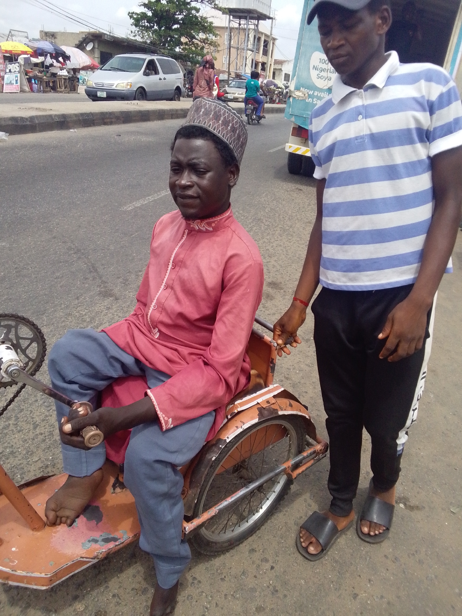 Two men are the primary subjects. One is sitting on a tricycle, wearing a pink shirt, blue pants, and a brown patterned cap. The tricycle appears to be homemade and painted orange. The man's bare feet are visible. The other man is standing next to the tricycle, wearing a blue and white striped polo shirt, black sweatpants, and black sandals. He is holding the tricycle.
The setting appears to be a street in Lokoja, Nigeria. In the background are cars, motorbikes, roadside stalls, and buildings. A truck with the words "100 Nigerian Soya" is partially visible in the background. The scene is captured in daylight.