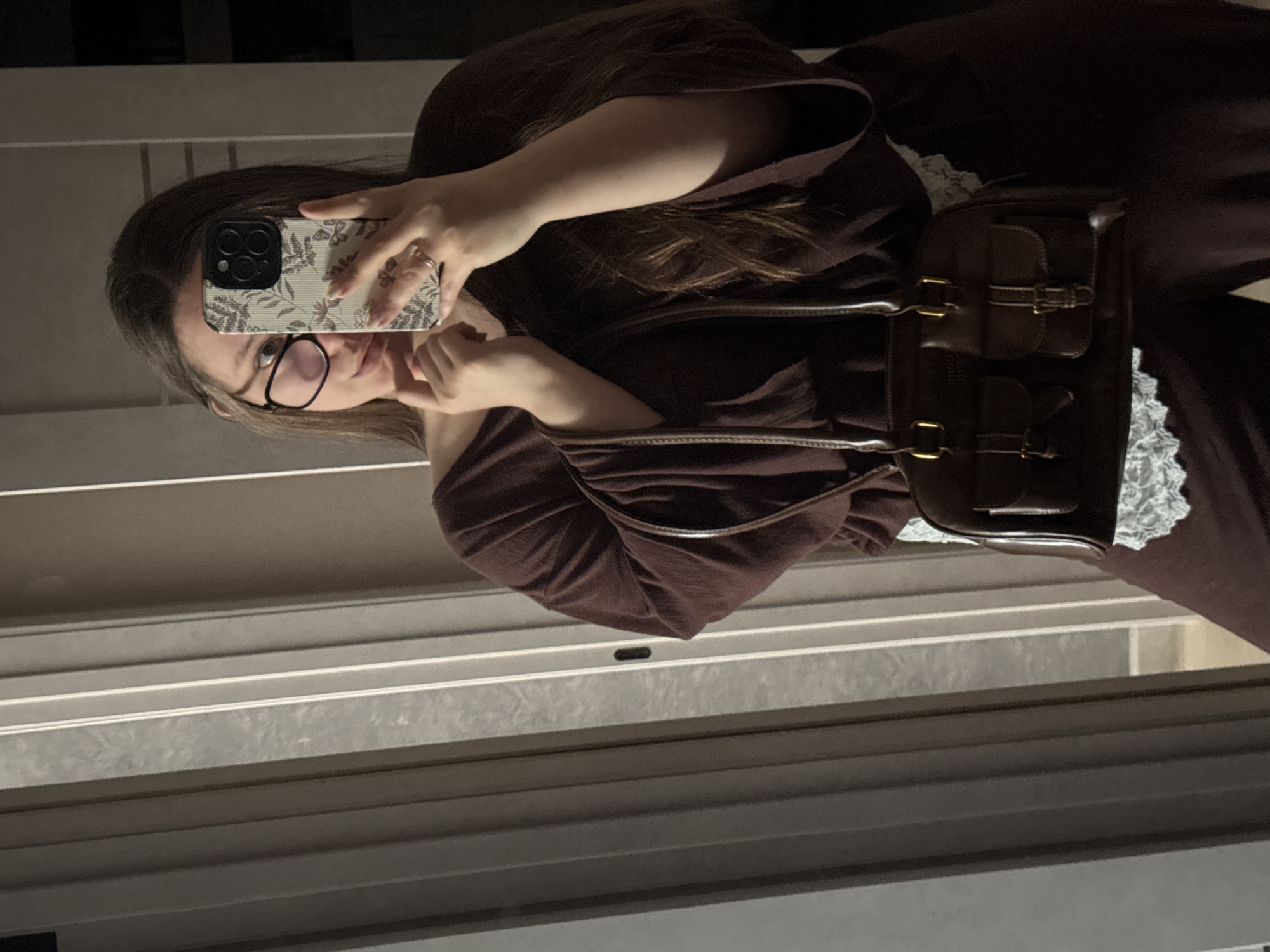 A young woman is taking a selfie in what appears to be a hallway or near a window. The woman is wearing glasses and a dark brown outfit with what may be lace trim. She is holding a cell phone with a patterned case, and the phone covers part of her face. She is also wearing a brown bag with buckle details. The background consists of a wall or window with horizontal lines. There is no information to confidently suggest this is in Krasnodar, Russia.