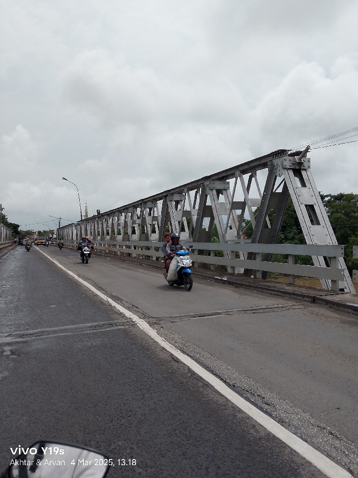 A view from the road on the bridge looking forward towards the other side of the bridge, in Jatibarang, Indonesia at coordinates -6.469 latitude and 108.297 longitude. The bridge is a metal truss structure. There is a single white line down the center of the asphalt roadway. Four motorcycles are visible travelling across the bridge. One motorcycle is closest to the viewer and contains two people. The sky is overcast. Trees are visible on the right side of the frame. The image was taken on March 4, 2025, at 1:18 PM. A mobile phone camera is visible in the lower left corner of the frame.