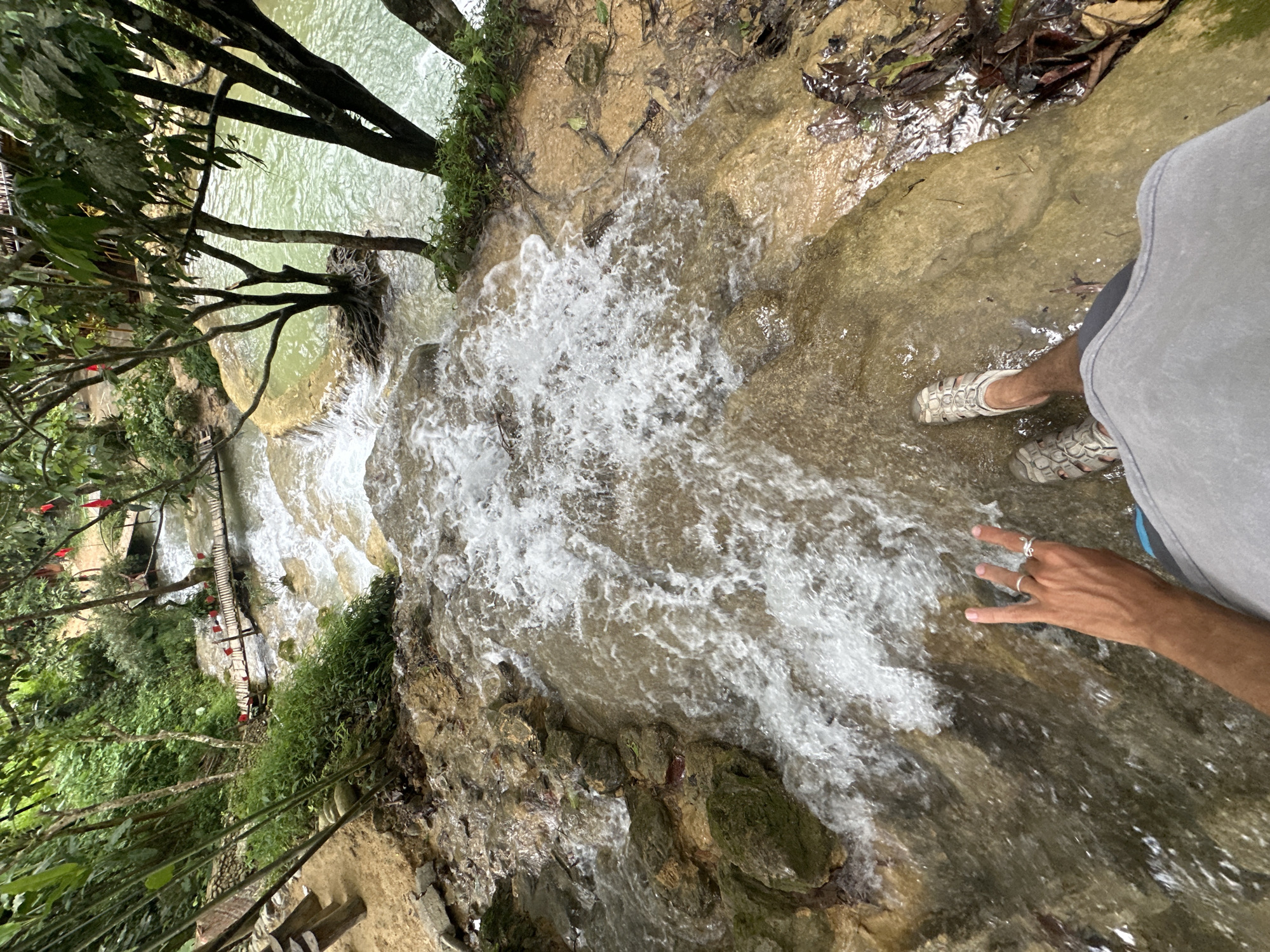 The image captures a person standing on a rock ledge overlooking a cascading waterfall in Cổ Lũng Commune, Vietnam. The person, partially visible, is wearing sandals and shorts. Their right hand is raised, with two fingers extended in a peace sign, and they are wearing rings on two fingers. The waterfall flows over textured, rocky terrain with patches of moss and vegetation. In the background, a lush green forest surrounds the waterfall, with trees and dense foliage. A small wooden bridge is visible further down the stream. The water is clear and appears to be flowing into a larger body of water in the distance. The scene is natural and serene, showcasing the beauty of the Vietnamese landscape.