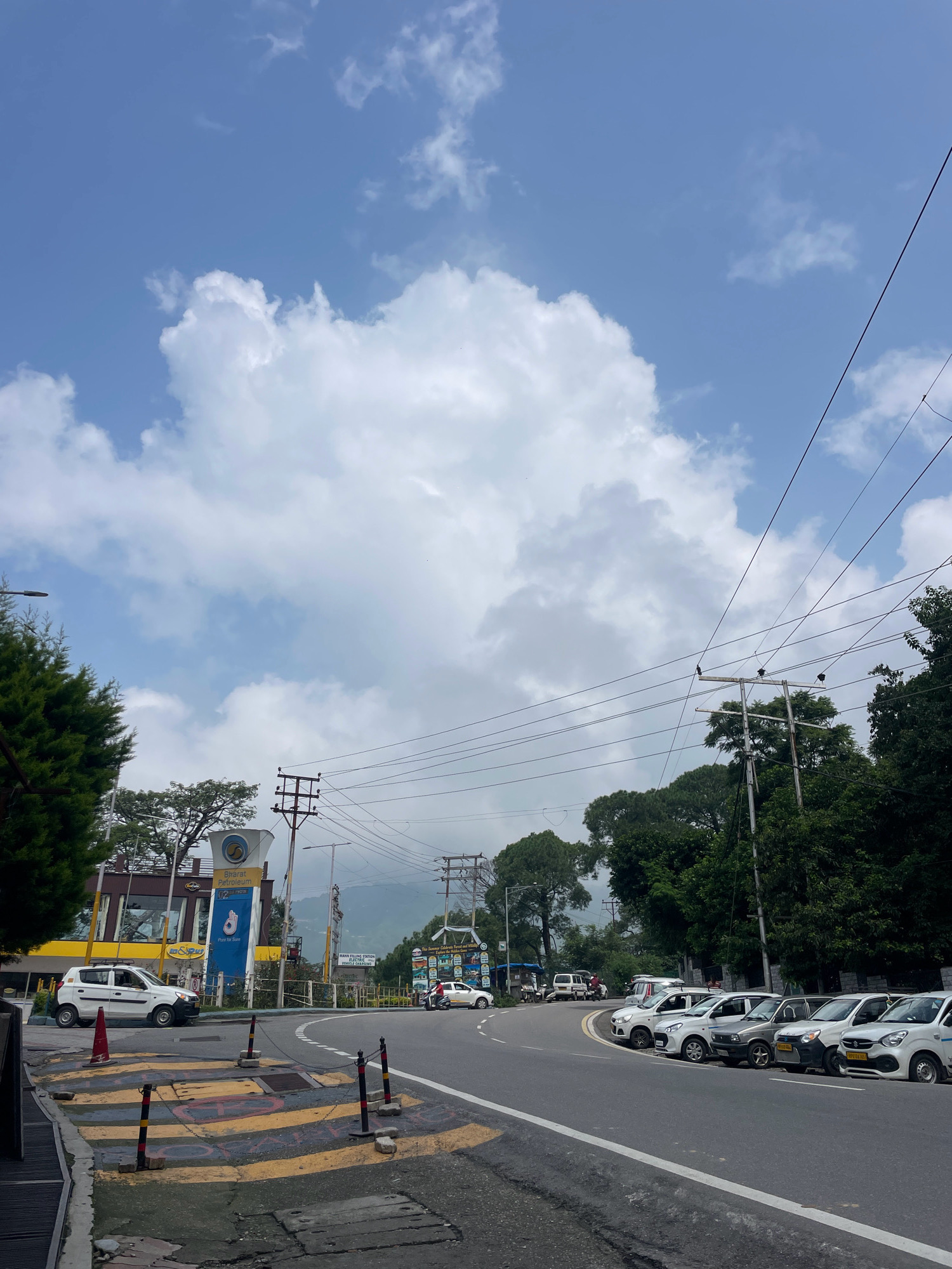 The image depicts a street scene in Dharamshala, India, likely on a cloudy day with the sky dominating the upper portion of the frame. A large white cloud formation is prominent against the blue sky. On the left side of the road, a white car is parked near a Bharat Petroleum gas station with a tall sign displaying the company logo and the word "Purity for Sure." Adjacent to the gas station, a dense green coniferous tree is visible.
The road curves to the right, with several other cars parked along its edge. These include a white hatchback, a grey hatchback, and other white cars. Power lines are strung across the scene, supported by utility poles, and a mountain range is visible in the distance, partially obscured by trees and clouds. The road surface appears to be asphalt, with white and yellow lane markings and a marked pedestrian crossing area with bollards. A few individuals are present near the gas station and on a motorcycle. The overall impression is that of a moderately busy road in a hilly, possibly urban or semi-urban area.