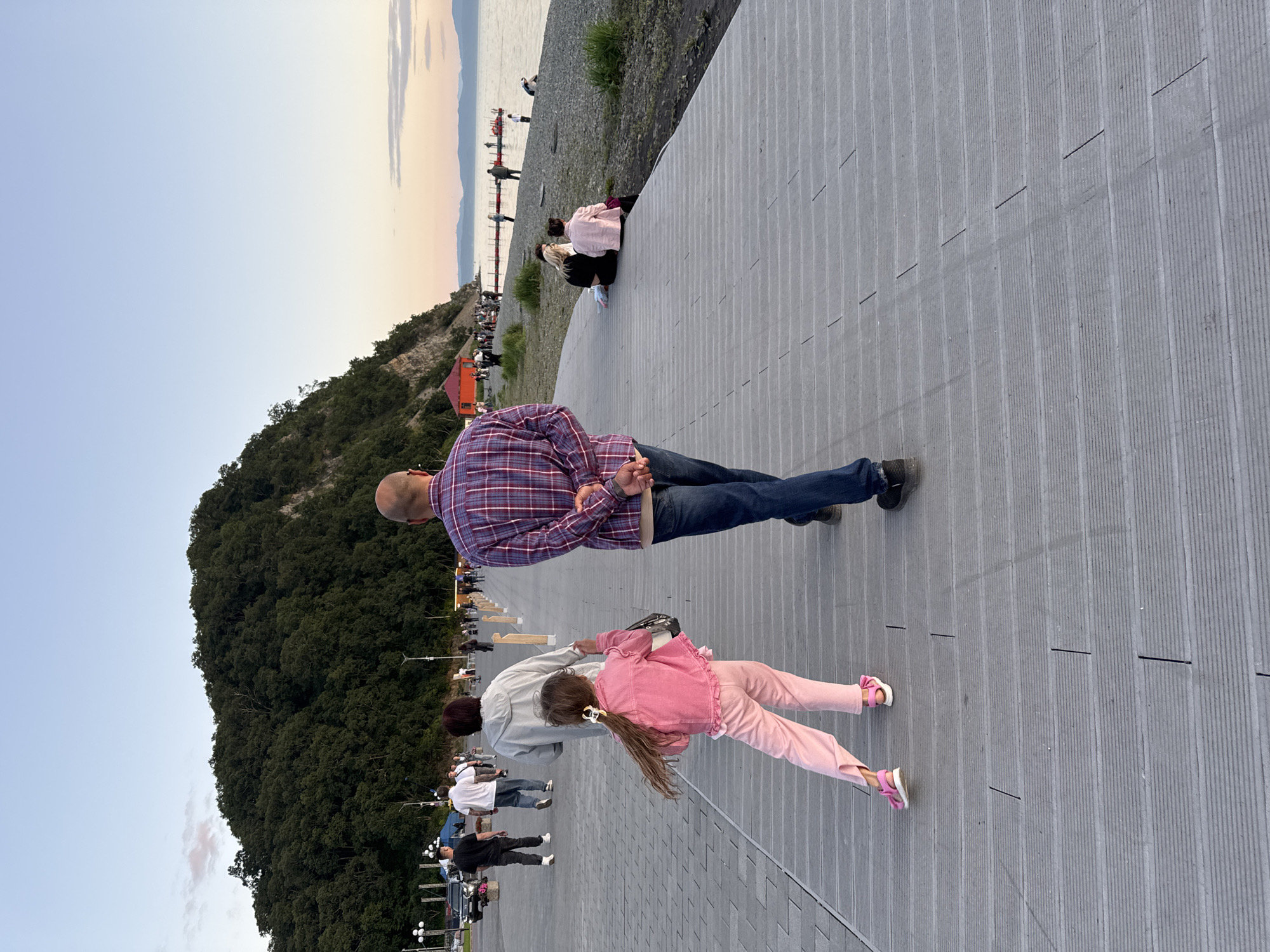 The image captures a promenade along a coastline, likely in Petropavlovsk-Kamchatsky, Russia, given the presence of a prominent, tree-covered hill characteristic of the region. The foreground shows a grey wooden boardwalk with a man and a young girl walking away from the camera. The man is wearing a plaid shirt and jeans, with his hands clasped behind his back. The girl, in front of him, is dressed in a pink jacket and pants, with her hair tied back with a yellow ribbon. She is looking back over her shoulder.
Further down the boardwalk, a woman in a grey hooded jacket walks with her back to the camera. In the background, other people are seen strolling, some sitting on the pebble beach, and a few are near the water's edge. A small red structure, possibly a tent or vendor stall, is visible near the base of the hill. The sea stretches out to the horizon, with faint hills visible in the distance. The sky is clear with a gradient of pale blue to soft orange, suggesting it is either sunrise or sunset. The overall scene depicts a relaxed evening atmosphere with people enjoying the seaside.