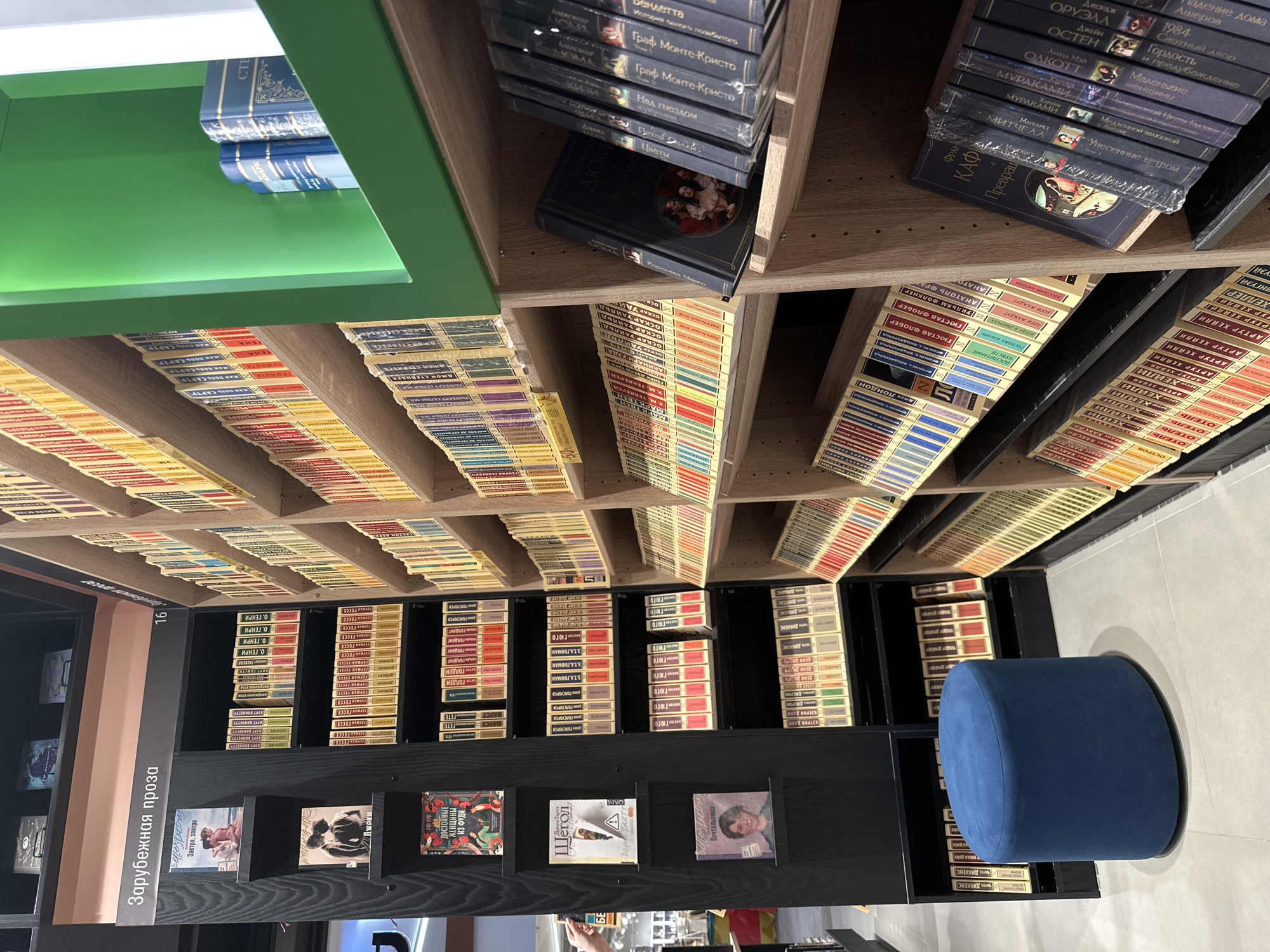 The image shows a section of a bookstore in Mukhanovo, Russia, featuring rows of books on shelves. On the left, a black bookshelf displays several books vertically, including titles with Russian text like "Зарубежная проза" (Foreign Prose). A blue ottoman sits in the foreground. To the right, wooden shelves are filled with numerous books, many with colorful spines. Some shelves are organized with books standing upright, while others are tilted. The overall impression is a well-stocked library or bookstore.