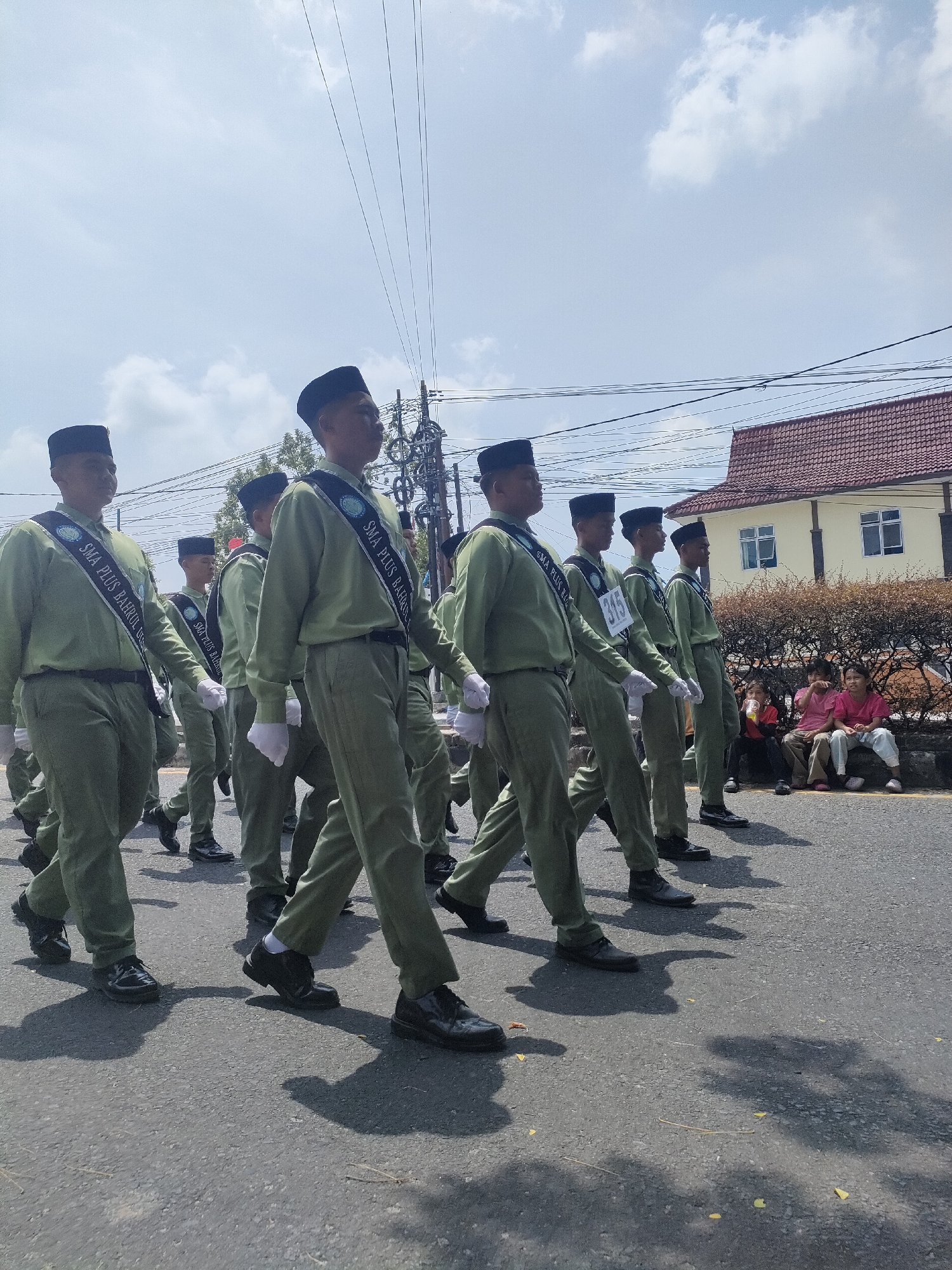 In Sungailiat, Indonesia, a group of young men, likely students, are marching in uniform. They are wearing light green long-sleeved shirts, matching trousers, black caps, and white gloves. A sash with the text "SMA PLUS BAHULUL" is worn across the chest of each individual. Some participants also wear bibs with numbers, such as "315." They are proceeding along an asphalt road, with power lines and a building with a red tiled roof visible in the background. Several bystanders, including children, are seated on a low wall to the right of the road, observing the parade. The sky is partially cloudy.