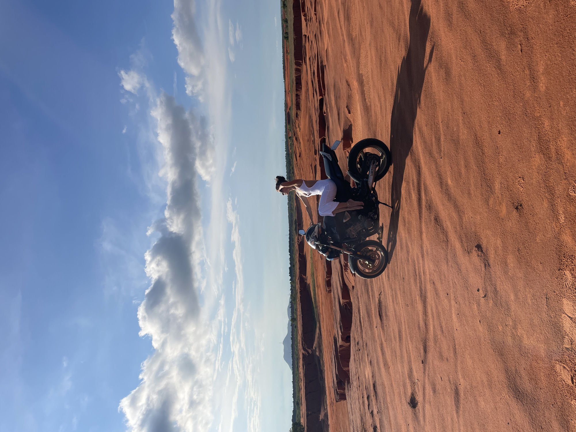 A young woman in a white dress is sitting on a black motorcycle. She is looking up at the sky, shielding her eyes with her hand. The motorcycle is parked on a red sand dune. In the background, there are more sand dunes and some red rock formations. A distant mountain is visible on the horizon under a blue sky with white clouds. This is likely the Red Sand Dunes of Mui Ne, Vietnam, a popular tourist destination known for its unique desert-like landscape.