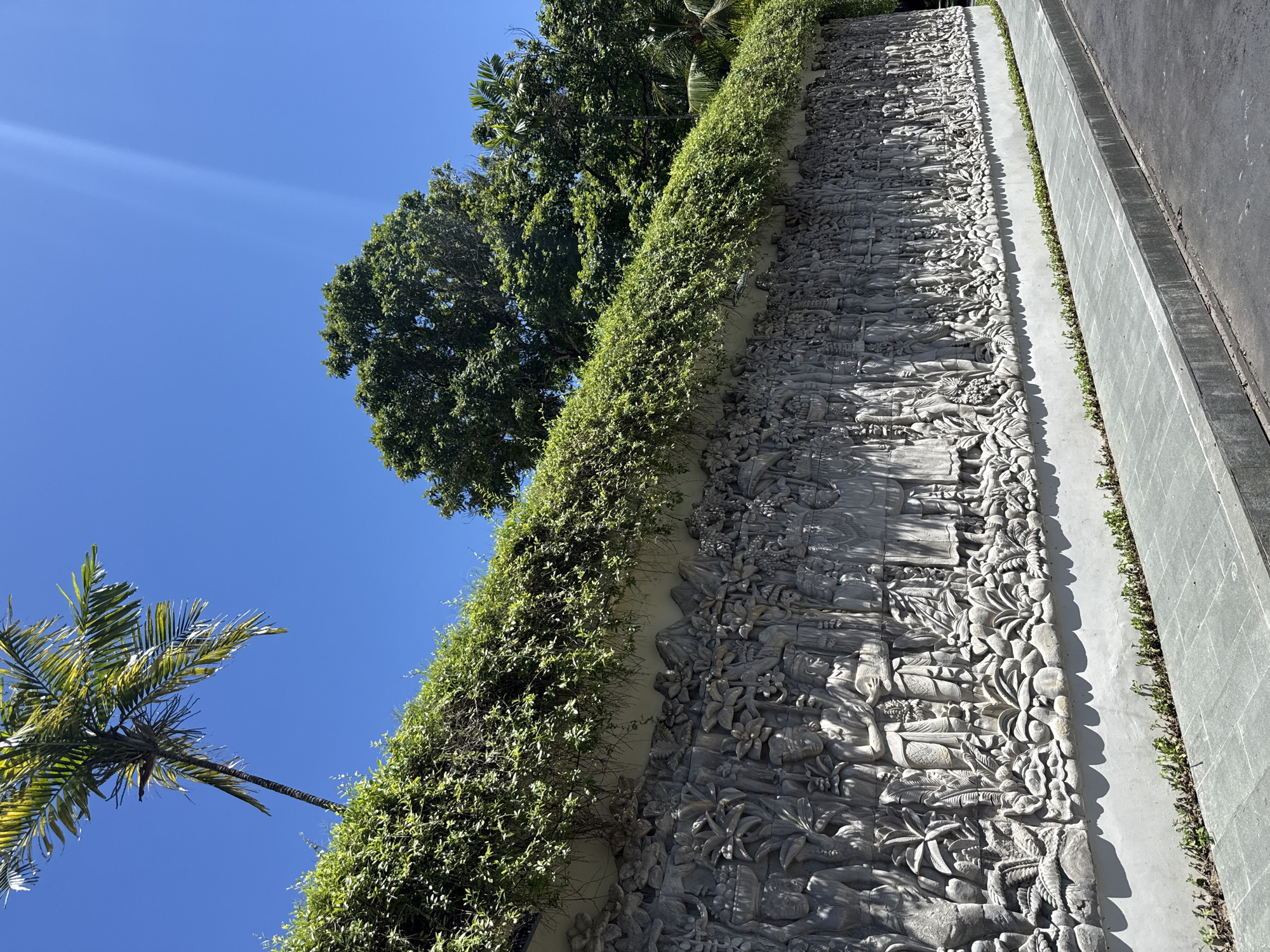 The image depicts a detailed stone carving wall in Nusa Dua, Indonesia. The wall features a complex relief depicting figures, plants, and scenes of daily life, partially obscured by a line of greenery growing atop it. To the left, the blue sky is visible, along with the tops of palm and leafy trees. A gray concrete structure borders the right side of the wall.