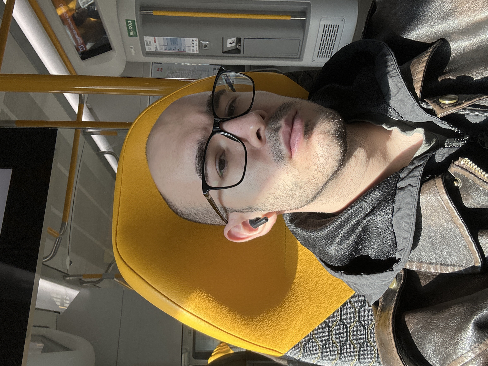 A male is seated in a public transit vehicle, possibly a bus or subway car, in Moscow, Russia. He is leaning back against a yellow headrest, wearing glasses and a black earbud in his left ear. The environment includes interior elements of the transit vehicle such as yellow handrails, a display screen, and overhead ventilation. He is also wearing a black mesh hoodie under a brown leather jacket.