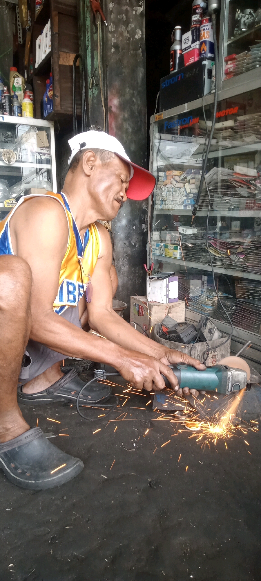 In Keteguhan, Indonesia, a man wearing a white cap with a red brim and a yellow and blue tank top is squatting down and using an angle grinder. Sparks are flying from the grinder as it cuts through metal. The man's legs are bare and he is wearing black slip-on shoes. The background shows shelves stocked with various items, possibly spare parts or tools, suggesting the location is a workshop or store. The ground is dark and appears to be dusty or dirty, with numerous sparks scattered across it. The lighting is dim, with the bright sparks from the grinder being the most prominent light source.