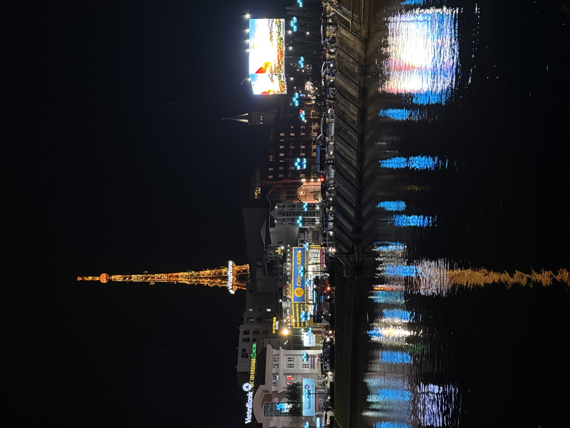 This media file depicts a nighttime scene of a riverfront city, with a prominent illuminated tower in the background. The scene is reflected in the dark water, creating abstract patterns of light and color. Buildings line the river, many adorned with neon signs and lights, suggesting an urban setting.
The illuminated tower, resembling a stylized Eiffel Tower, is a central object, decorated with a multitude of lights. Various buildings are visible, some appearing to be commercial establishments with signage. One large billboard or screen displays an image of a landscape with what appears to be a flag. The water reflects the city lights, with streaks of white, blue, and orange creating a dynamic visual effect.
The time of day is clearly night, with the dark sky contrasting with the bright lights. The weather appears calm, as the water is relatively still, allowing for clear reflections. The presence of numerous illuminated signs and the style of the architecture suggest this is an urban environment in Southeast Asia, consistent with the location context of Da Lat, Vietnam. While no specific event is discernible, the overall atmosphere is one of lively nighttime activity and urban illumination.
Visible text includes "Vinaphone," "VietinBank," "Wisteria Hotel," and "DIEN XANH."