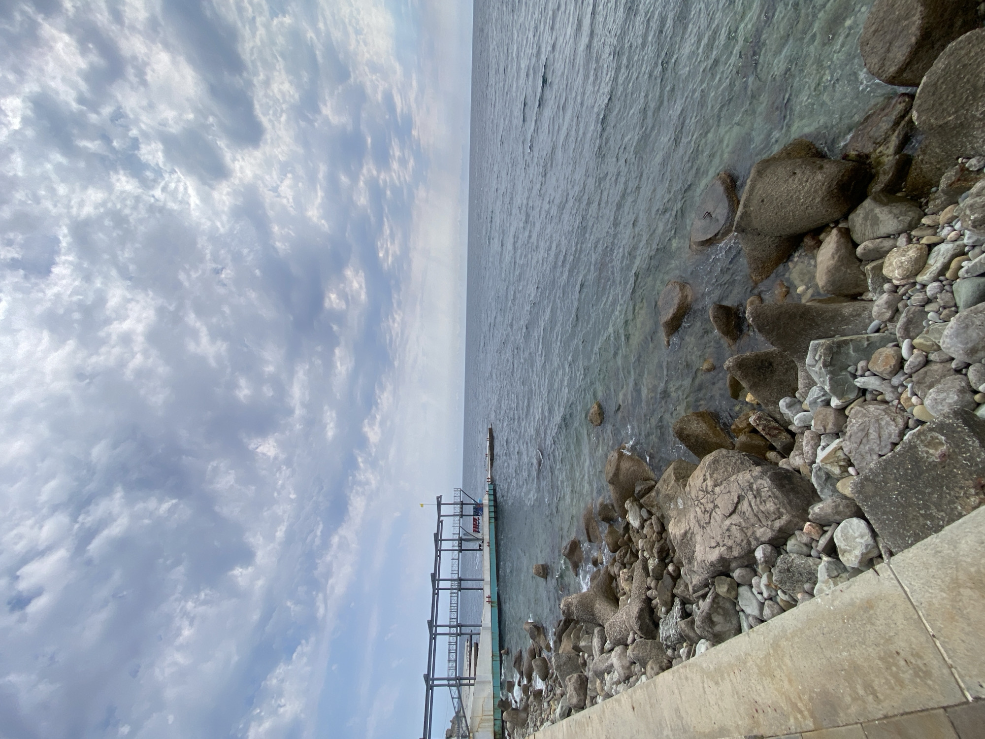 The image shows a view of the Black Sea from a rocky shoreline in the settlement of Gaspra, Yalta Municipality, Crimea, Ukraine. In the foreground, a concrete pathway lines the edge of the shore, which is covered with a variety of large and small stones and rocks. Some of these rocks appear to be tetrapods, used as a breakwater. The sea is calm with gentle ripples, and the water is clear enough to see submerged rocks in some areas. In the middle ground, a pier or structure extends out over the water, supported by metal beams and a turquoise-colored base. There is a yellow flag visible at the end of this structure. The horizon line where the sea meets the sky is visible in the distance. The sky is overcast with scattered clouds. No people are visible in the frame.