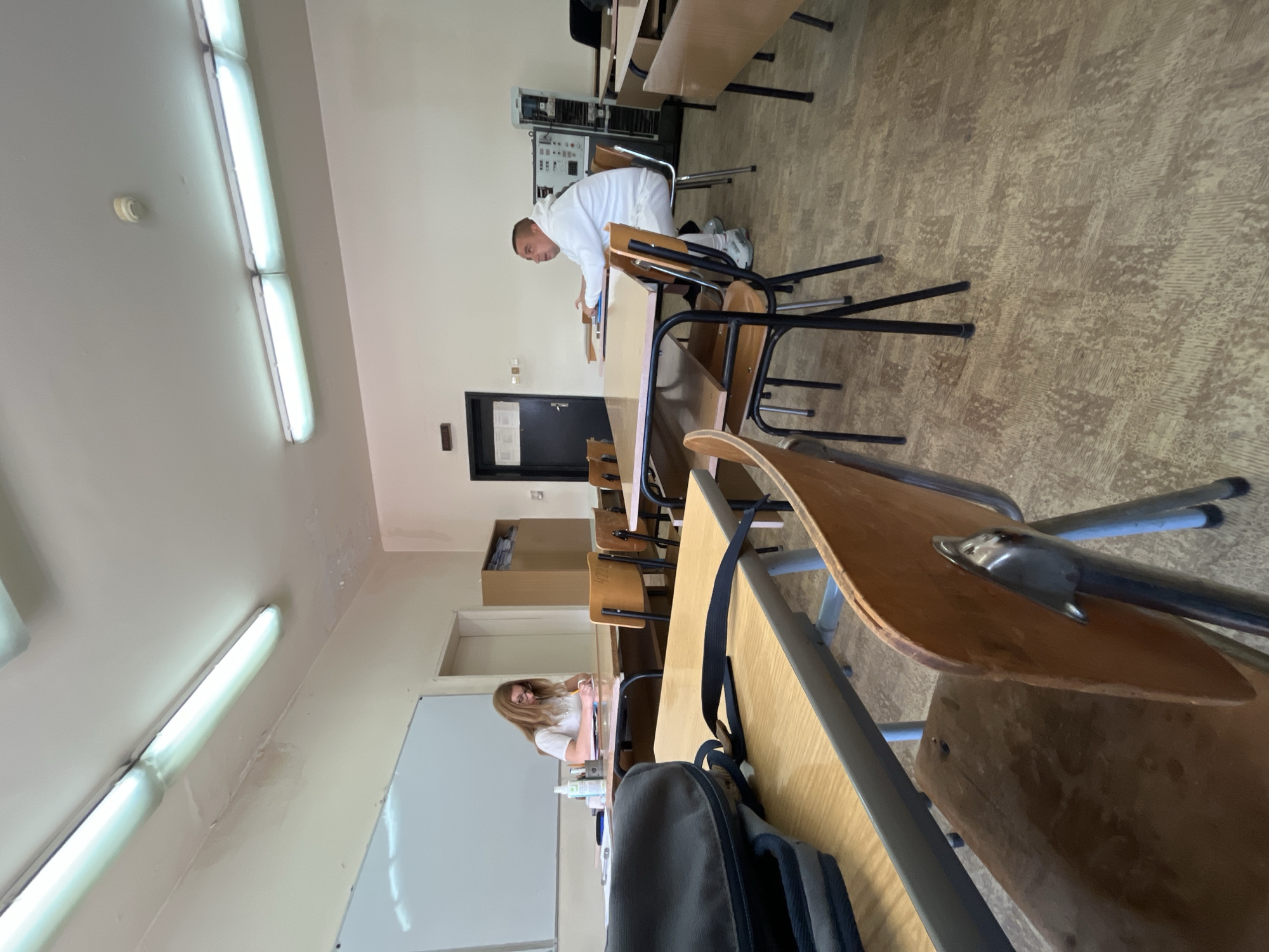 **Location:** Classroom, likely in an educational institution, Plovdiv, Bulgaria.
**Environment:** The classroom has fluorescent tube lighting and tiled flooring. Multiple rows of desks and chairs face forward. A whiteboard is visible on the left side of the frame.
**People:** Two individuals are present. A man in a white tracksuit is leaning over a desk. A woman with long hair wearing a white shirt sits at a desk towards the front of the room, writing.
**Objects:** Desks, chairs, a backpack, and writing utensils.
**Actions and Interactions:** One person is writing at a desk, while another appears to be reaching for or interacting with something on a different desk.