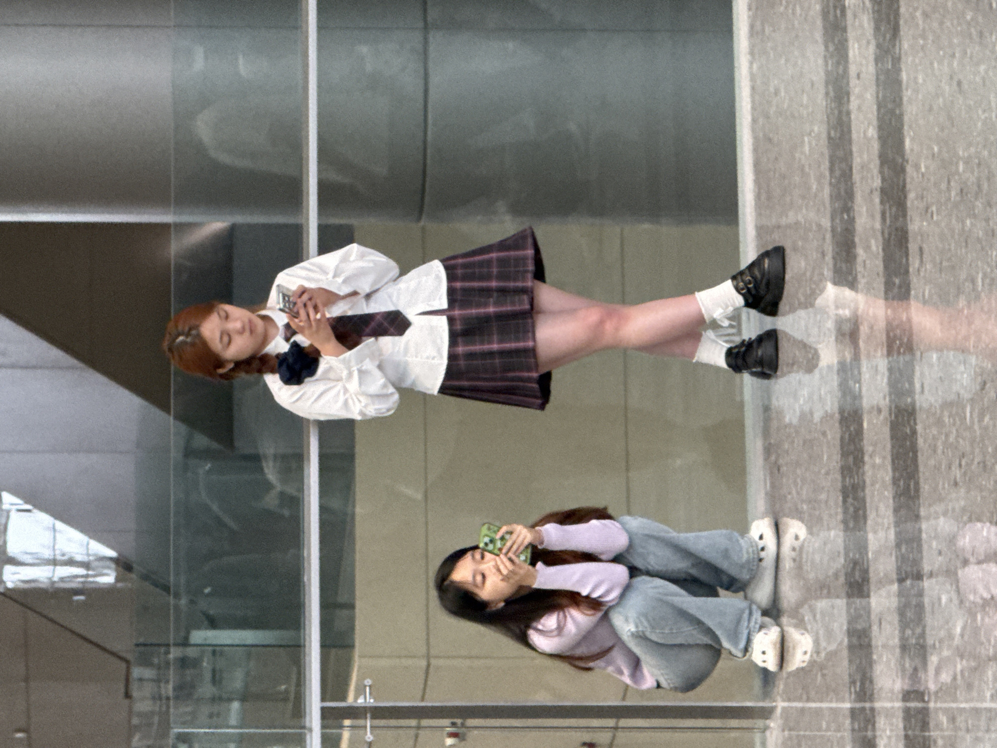 Two young women are standing in a modern, indoor space with polished floors and large windows. The woman on the left is crouching and looking at her phone, which is held in her hands. She is wearing a light purple sweater and light blue jeans, along with white clogs. Her hair is dark and falls around her shoulders. The woman on the right is standing and also looking at her phone. She is wearing a white button-up shirt, a plaid skirt, and dark shoes with white socks. Her hair is reddish-brown and styled in braids with a dark bow. Behind the women, large pillars and glass partitions suggest an office building or public space in a city. The overall lighting is bright, reflecting off the shiny floor. The scene appears to be a public area within a commercial or educational institution in Chongqing High-tech Zone, China, possibly during a break or while waiting.