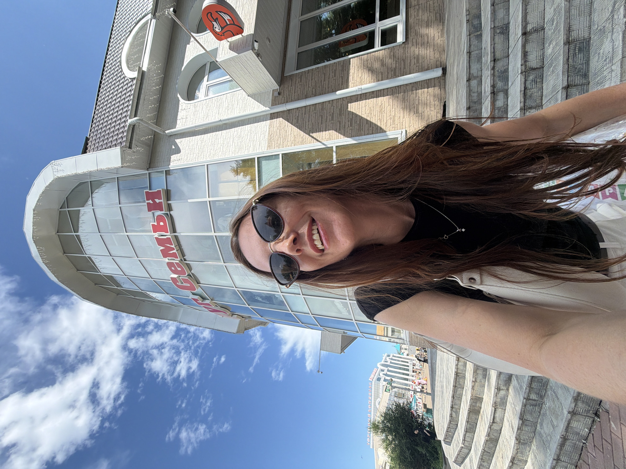 A woman with long brown hair and sunglasses smiles at the camera while holding a white bag, in front of a building with a curved glass facade. The building has red lettering on its facade. The woman is standing on a set of concrete stairs. In the background, there is a clear blue sky with scattered clouds, and other buildings and people are visible. The scene appears to be in the city of Birobidzhan, Russia.