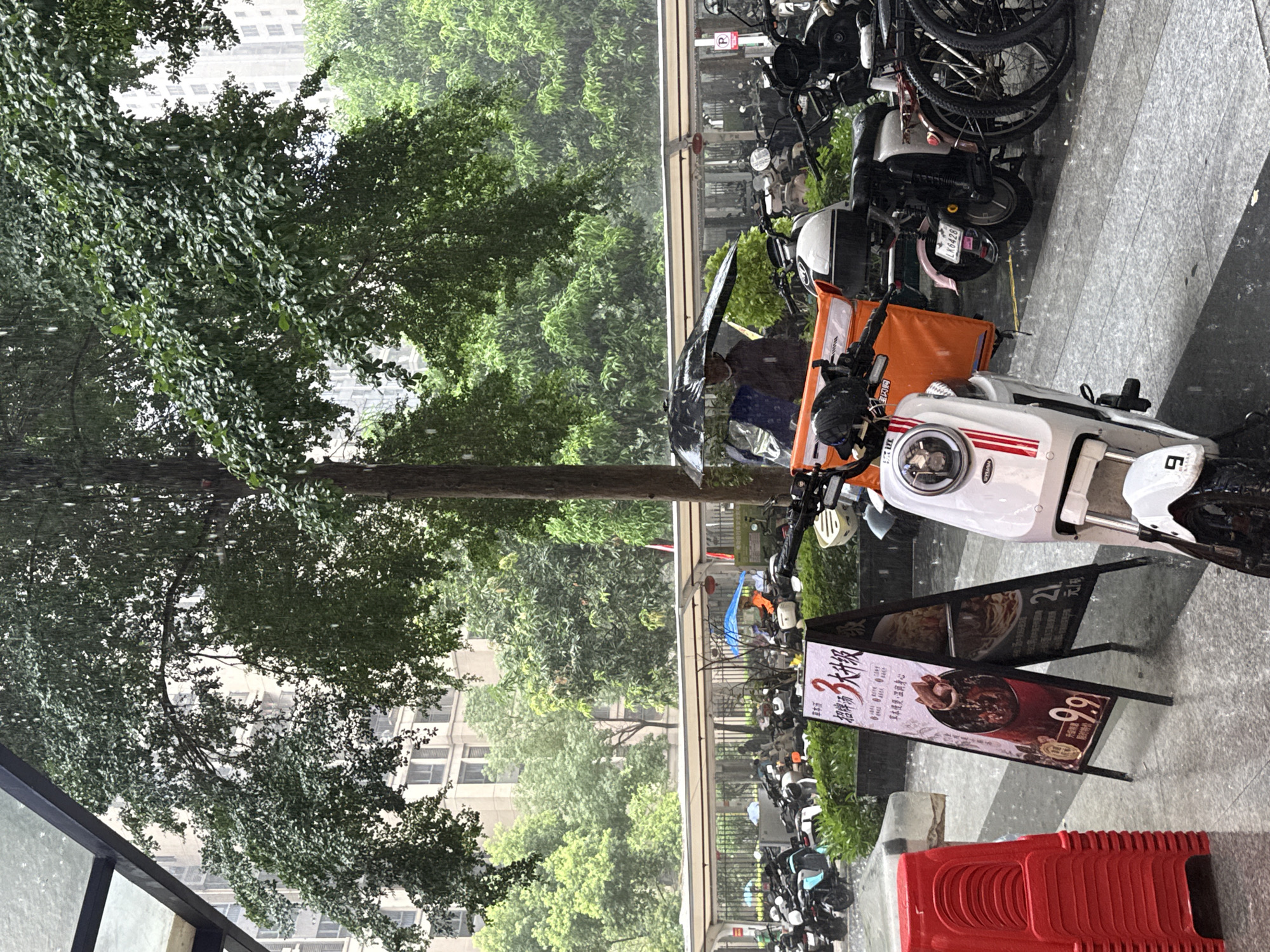 The image captures a street scene in Haizhu District, China, during a rain shower. The focus is on a row of parked electric scooters and bicycles. One scooter in the foreground is white with red and black accents and has a license plate that reads "LK6428". An orange delivery bag is attached to another scooter. A-frame signs are placed on the wet pavement, advertising food with prices like "9.9" and "21". One sign features an image of a dish with meat. In the background, lush green trees are visible, with the suggestion of buildings beyond. The rain is evident through the blurriness of the foliage and the wet appearance of the ground and vehicles. A person holding a black umbrella is partially visible, suggesting they are either preparing to use it or have just taken shelter. Red plastic chairs are stacked neatly to the side. The overall atmosphere is one of a typical urban day disrupted by rain, with delivery services and street-level commerce continuing amidst the downpour. The text visible on the signs is in Chinese, mentioning "草本慢煲" (herbal slow-cooked) and "招牌大升级" (signature dish upgrade).