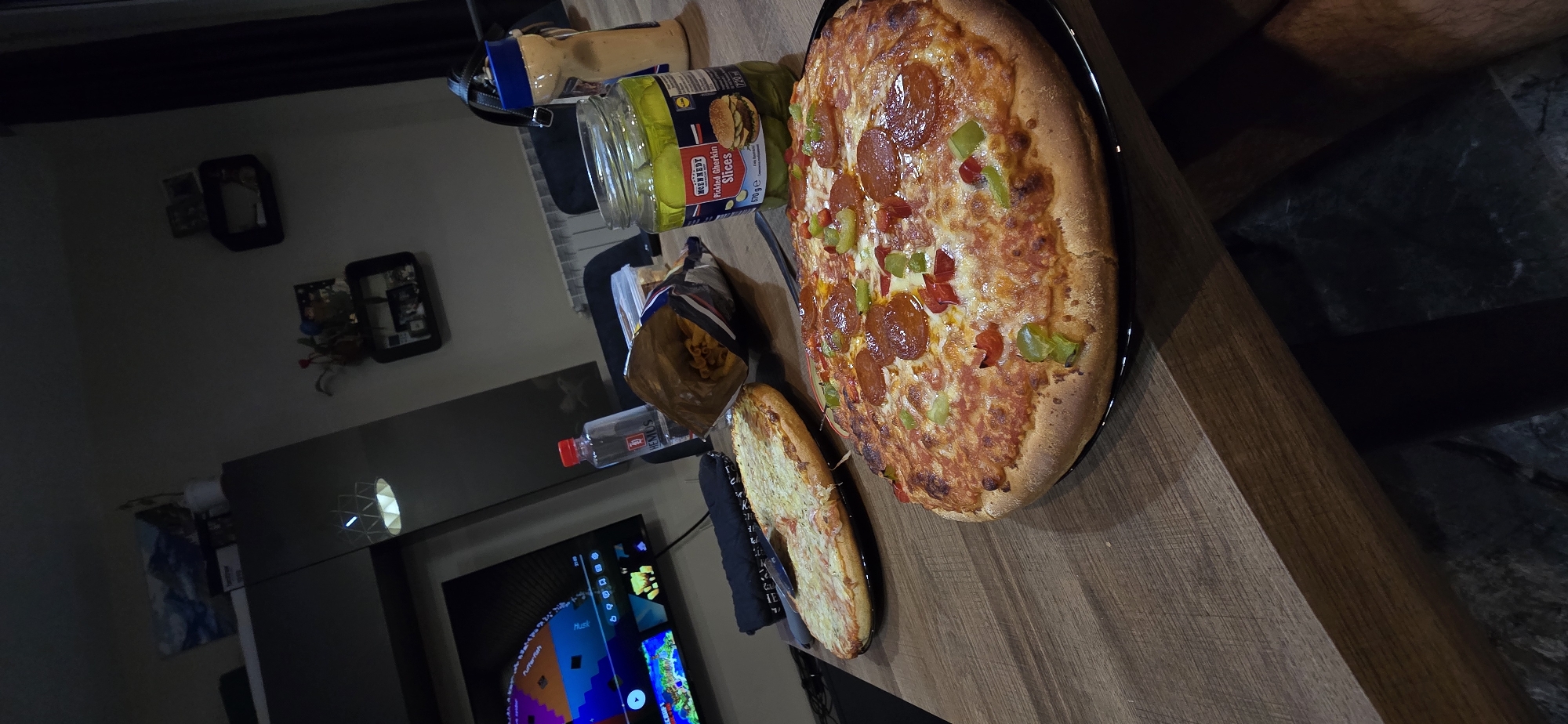 The image depicts a dimly lit indoor scene, likely a living room or dining area. A wooden table occupies the foreground, laden with food items and drinks. Two pizzas are prominently displayed: one with what appears to be cheese and possibly white sauce, and another topped with pepperoni, green peppers, and red peppers. To the right of the pizzas, a jar of "McEnnedy Pickled Gherkin Slices" is visible. Next to the jar is a bottle of water with a red cap and a partially open bag of snacks, possibly chips or pasta. A light-colored bottle, possibly a condiment or dressing, is also present. In the background, a television screen displays colorful graphics. The room's ambiance is dark, with artificial lighting creating shadows and highlighting the objects on the table. There are no people visible in the image. The location is not specified in the image.