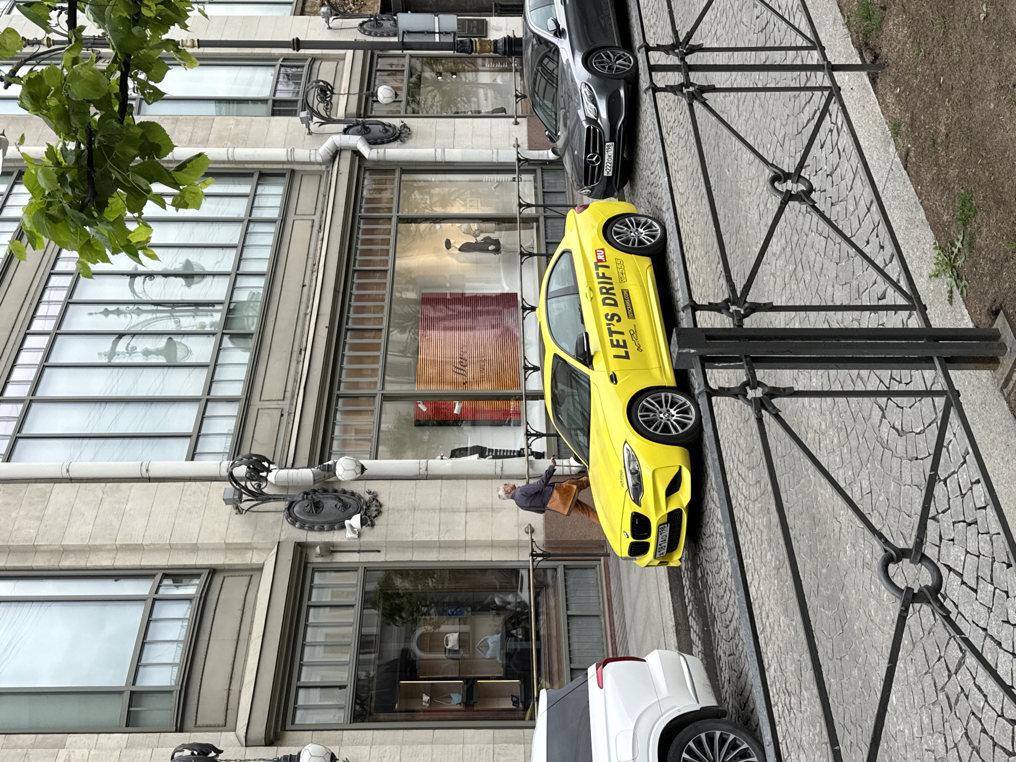 A man walks past a bright yellow BMW M2 coupe parked on a cobblestone street in front of a multi-story building with large windows. The building appears to be a retail establishment with mannequins visible in the window displays. The BMW has a sticker on its side that reads "LET'S DRIFT.RU". A dark-colored Mercedes-Benz E-Class is parked behind the BMW. The scene is on a street in Saint Petersburg, Russia.