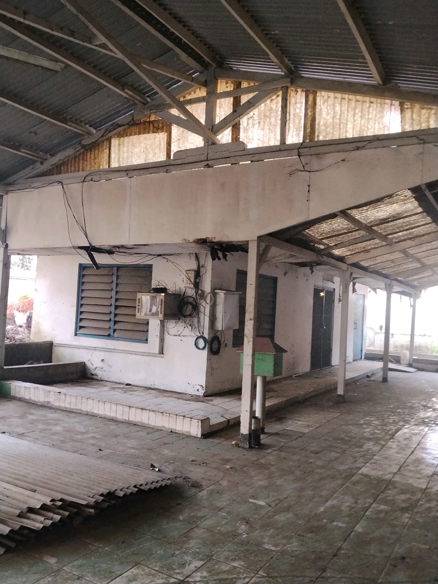 The image depicts an abandoned, partially dilapidated structure, possibly an old warehouse or a community building, located in Suka Nagara, Indonesia. The building features a corrugated metal roof supported by wooden beams. The exterior walls are a weathered white, with visible signs of age and neglect. Several windows are present, some with louvers that are closed. An air conditioning unit is mounted on one of the walls. Electrical wiring is exposed and hangs haphazardly. The ground in front of the building is paved with tiles that appear to be stained and worn. A green post box-like structure stands near the entrance. The overall scene conveys a sense of disuse and decay.