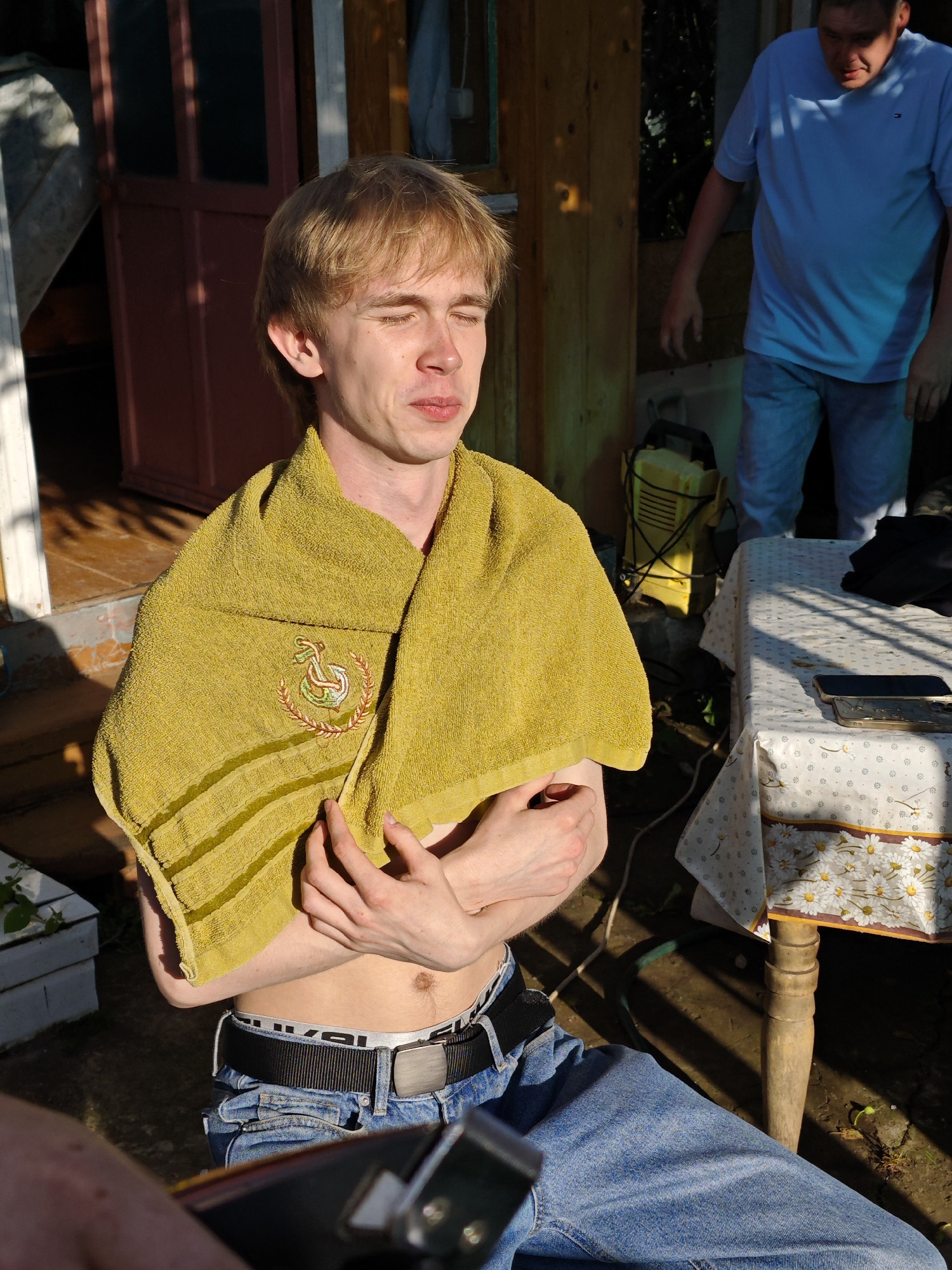 The image depicts two people in an outdoor setting, likely a backyard or a rural area in Foky, Russia, as suggested by the architectural style of the building in the background and the presence of a wooden table. The time of day appears to be late afternoon or early evening, indicated by the warm, directional sunlight casting long shadows and highlighting the subjects.
The primary subject is a young man with blonde hair, sitting in the foreground. He is wearing blue jeans and a towel draped over his shoulders. The towel is a light yellow-green color and has an embroidered emblem of an anchor within a laurel wreath. The man has his eyes closed and appears relaxed, possibly enjoying the warmth of the sun or contemplating something. His arms are crossed, and he is leaning slightly forward.
In the background, another person, a man wearing a blue t-shirt and jeans, is standing and looking down. He appears to be in the process of setting up or operating some equipment, possibly related to yard work or maintenance, as a yellow generator or power tool is visible near him. A wooden table covered with a patterned tablecloth stands between the two individuals, with a few objects on it, including what appear to be mobile phones.
The overall atmosphere is casual and relaxed, suggesting a summer day. The scene captures a candid moment, with the sunlight creating a natural and somewhat rustic ambiance. There is no visible text in the image.
