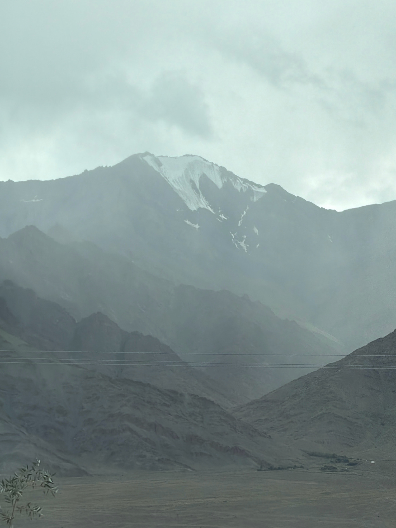 This is a vertical, medium-distance, outdoor, eye-level shot of a hazy, mountainous landscape. The sky is overcast and pale, with diffused light. In the background, a large mountain range dominates the scene. The peak of the highest mountain is capped with snow, with streaks of white snow running down its grey, rocky slopes. In front of this, layers of smaller, darker, undulating mountains recede into the haze. Two parallel wires are stretched across the middle ground, supported by unseen poles. The foreground is a muted brown, flat, open plain, with some low, scrubby vegetation visible on the left. In the far distance, on the right side of the plain, there appear to be a few small structures and a cluster of trees. The overall impression is one of a vast, rugged, and somewhat desolate natural environment under a muted sky. The scene appears to be located in the region of Yuluk, India, given the style of the landscape and the presence of what appear to be power lines.