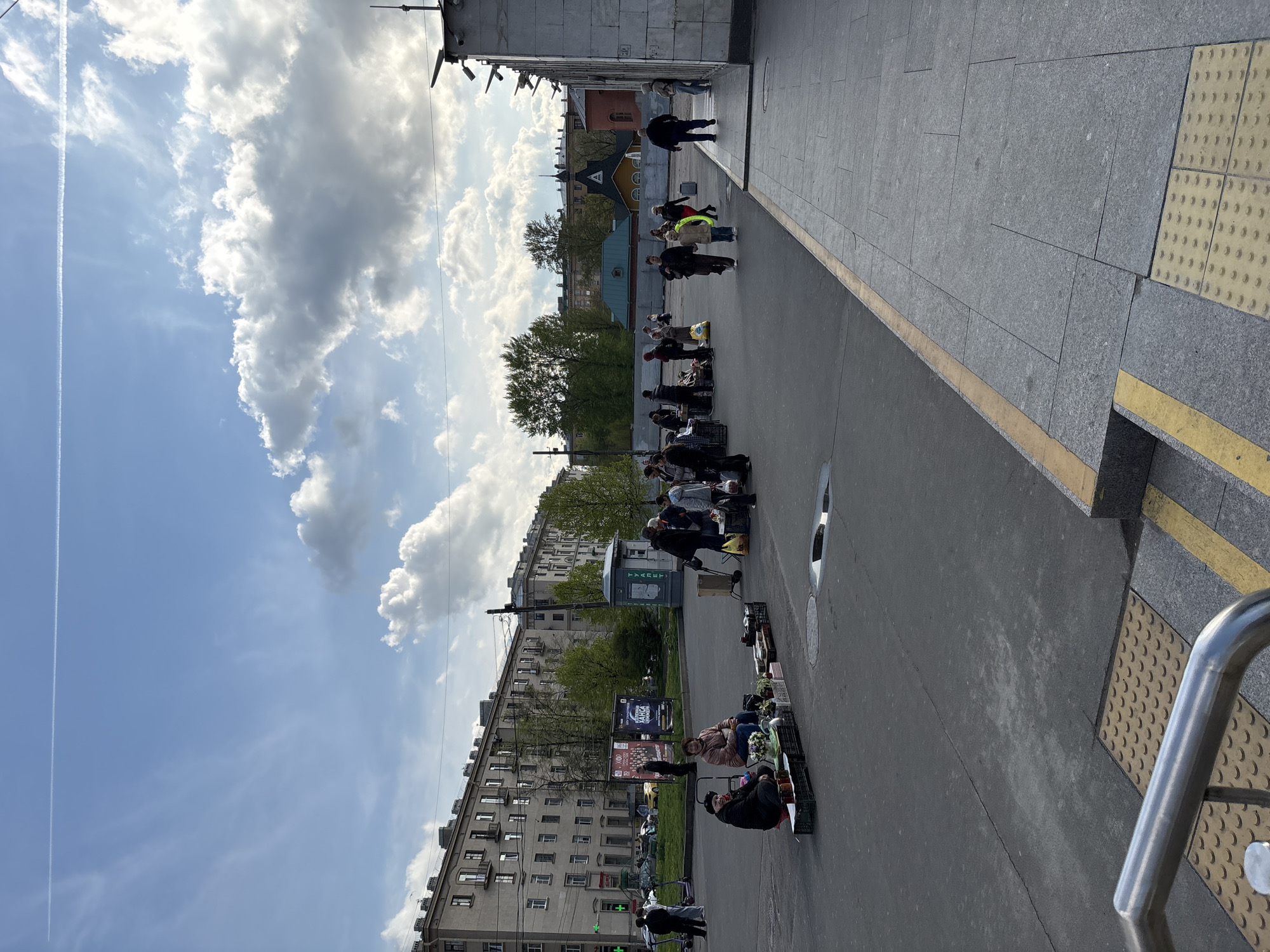 The image depicts a street scene in Saint Petersburg, Russia, likely taken on a sunny day with scattered clouds. A queue of people stretches along the sidewalk adjacent to a busy street. Some individuals are seated, possibly vendors, with various items displayed for sale, including flowers and other merchandise on pallets. Buildings line the opposite side of the street. The sidewalk is paved with gray stone, and a yellow tactile strip indicates a curb edge. A metal handrail is visible in the foreground. In the background, are apartment buildings, street signs, and green trees. The sky is bright blue with prominent white clouds and a contrail. The overall impression is of a typical urban scene with commercial activity and pedestrian traffic.