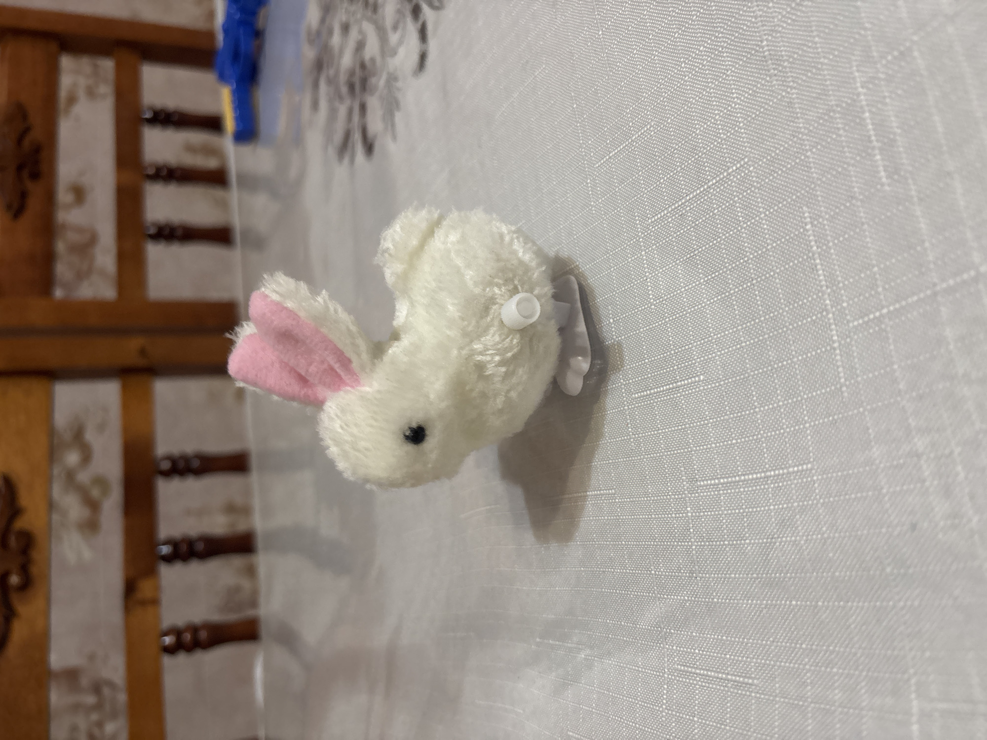 The image captures a small, fluffy white toy rabbit resting horizontally on a light-colored, textured fabric surface, possibly a tablecloth or bedspread. The rabbit features soft white fur, distinct pink inner ears, and a small black nose. A white plastic wind-up key is visible on its right side, indicating it is a wind-up toy. A small white appendage, likely a tail or foot, extends from its rear.
In the blurred background on the left, a segment of a brown wooden structure with decorative elements, possibly part of a furniture piece like a bed frame, is visible. Behind this wooden element, a lighter surface with faint patterns can be discerned. A blue object is partially visible in the upper left corner. The textured fabric beneath the rabbit exhibits a fine grid-like weave.
The setting is an indoor environment, suggesting a domestic space. No specific activity is occurring; the toy is static. The lighting indicates an indoor scene, but the precise time of day is not discernible. No text is visible in the media file. The location context provided is Ферма, Russia.