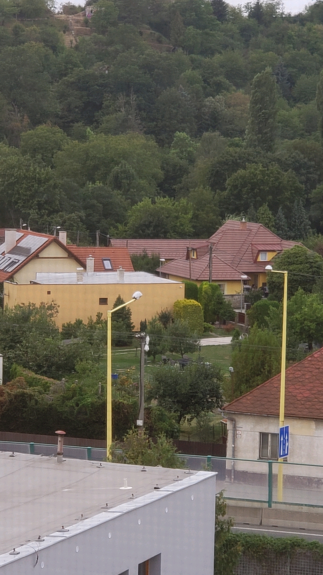 The image shows a residential area with houses and trees in the city of Prešov, Slovakia. In the foreground, a flat-roofed building is visible, with a grey facade and a white roof. Further back, a yellow house with a red tiled roof is prominent, featuring several windows and a skylight. Adjacent to this yellow house is another building with a red tiled roof and yellow trim. The scene is surrounded by dense green trees, suggesting a semi-urban or suburban environment. Two yellow lampposts are visible in the middle ground, one on the left and one on the right, with a pedestrian crossing sign on the right. The sky is overcast.