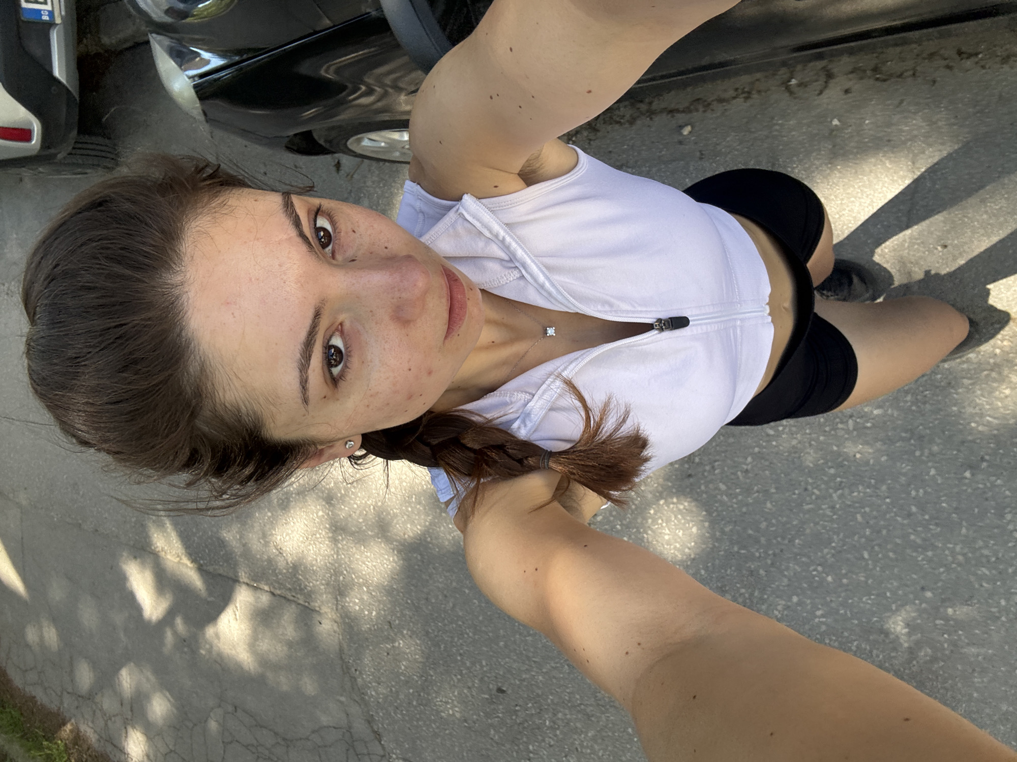 A woman in athletic attire stands on a paved surface next to a car. She is wearing a white zip-up tank top, black shorts, and athletic shoes. Her brown hair is in a braid. A diamond pendant necklace is visible on her chest. The car is a dark color, possibly black. The pavement is grey. Tree shadows are cast on the ground. The image is a selfie taken from a high angle. There is no specific information available to determine if the photo was taken in Varna, Bulgaria.