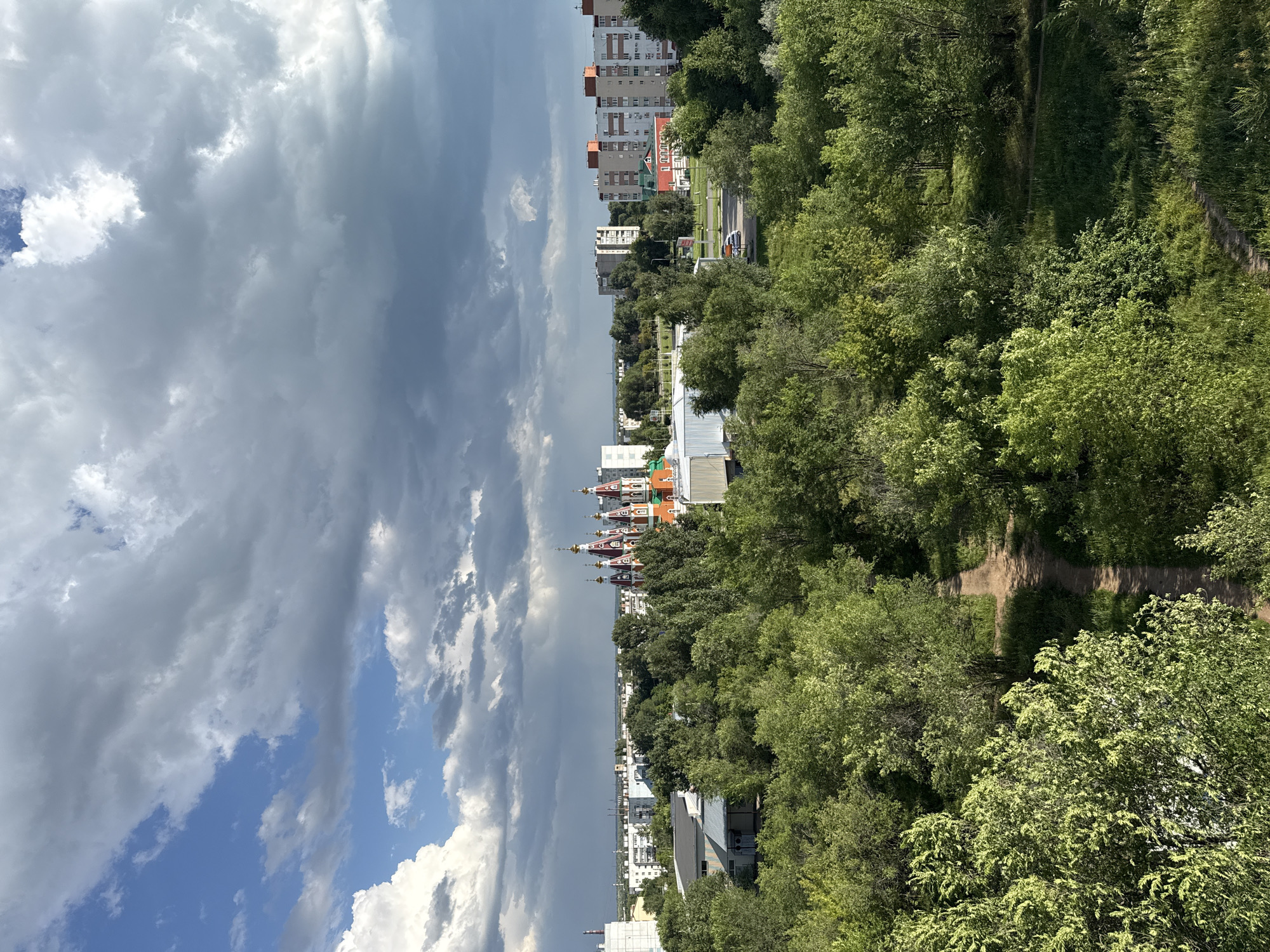 The image depicts a view of the city of Otradny, Russia, with a prominent red and white church featuring golden domes in the middle ground. The foreground is filled with lush green trees and a dirt path winding through them. Behind the trees, the church stands out against a backdrop of residential buildings and a cloudy sky. Some apartment buildings with red accents are visible to the right of the church. A road with a white car is also present. The sky is a mix of blue patches and heavy grey clouds, suggesting a potentially changeable weather condition.