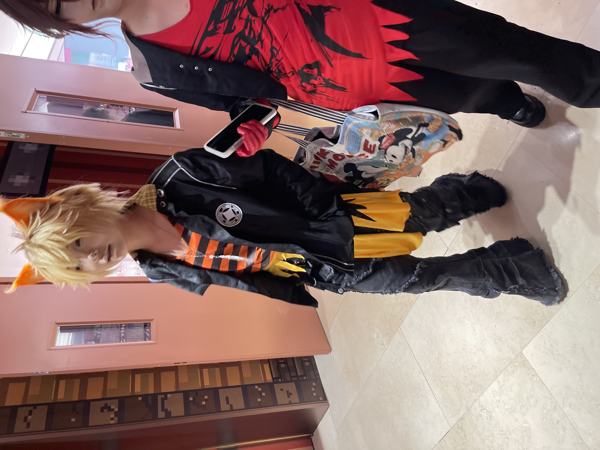 In this image, two individuals are depicted, likely at an indoor event or gathering. The person in the foreground, with blonde hair and orange fox-like ears, is wearing a black jacket with orange and black striped sleeves, and black pants. They are holding a guitar with a Mickey Mouse themed design. To the left of this individual is another person wearing a red top with black accents and black pants.
The scene appears to be indoors, with a tiled floor and wooden paneling visible in the background, suggesting a public space or venue. The lighting is artificial and evenly distributed, indicating it is not daytime or an outdoor setting. No specific emotions are discernible from the subjects' expressions. The overall impression is of a casual or themed event, possibly a cosplay or costume party, given the attire. There is no visible text in the image.