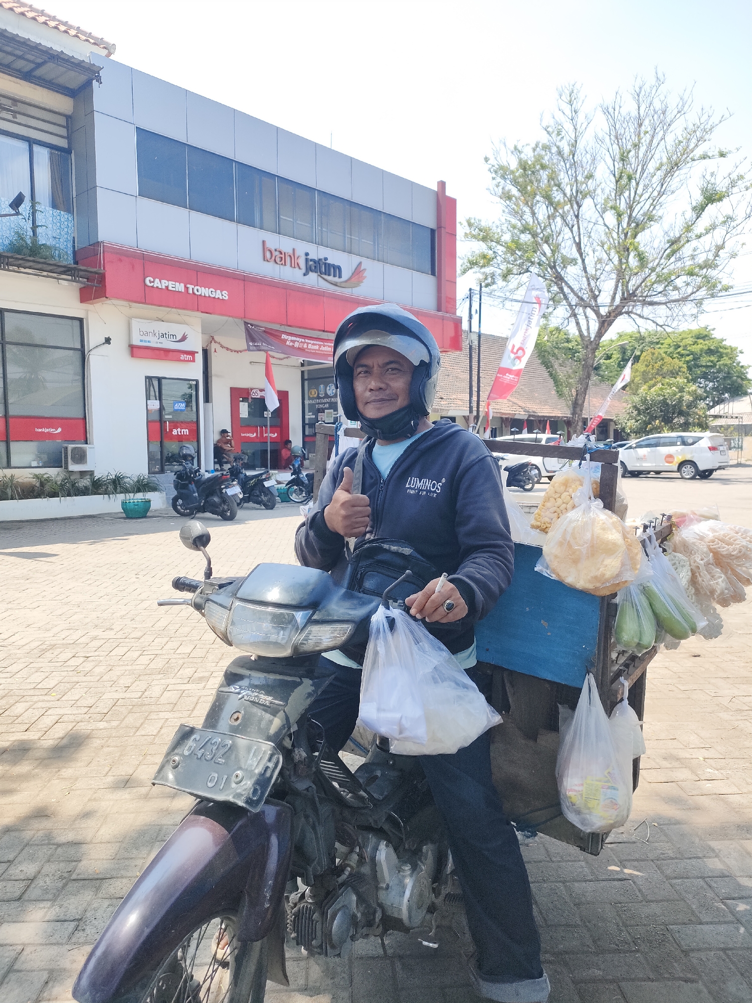 In Curahdringu, Indonesia, a man wearing a helmet and a dark blue hoodie with the logo "LUMINOS" gives a thumbs up while sitting on a motorcycle. The motorcycle has a license plate that reads "6432 WH 01 20." Attached to the back of the motorcycle is a blue cart carrying several plastic bags filled with food items, including what appears to be crackers and possibly vegetables. In the background, the "Bank Jatim" building with "CAPEM TONGAS" signage is visible. Several other motorcycles and a white car are also present in the scene, along with trees and flags. The ground is paved with interlocking bricks. The overall scene depicts a sunny day in an urban environment.