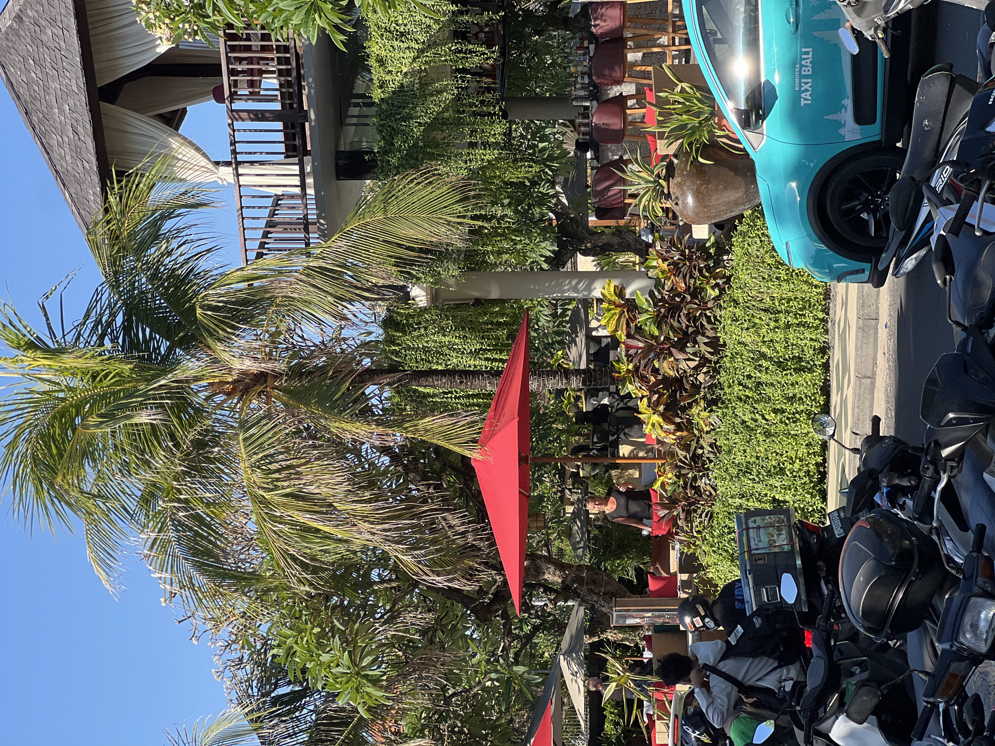 In Kuta, Indonesia, a bright blue Komotra taxi, branded "TAXI BALI," is parked on a street. In the foreground, several motorcycles are parked, some with riders preparing to depart. Behind the motorcycles, a woman in a black top stands near a red umbrella, amidst lush green foliage and outdoor seating areas with red cushions. A multi-story building with a balcony adorned with white curtains and a dark wooden railing looms in the background, partially obscured by palm trees.