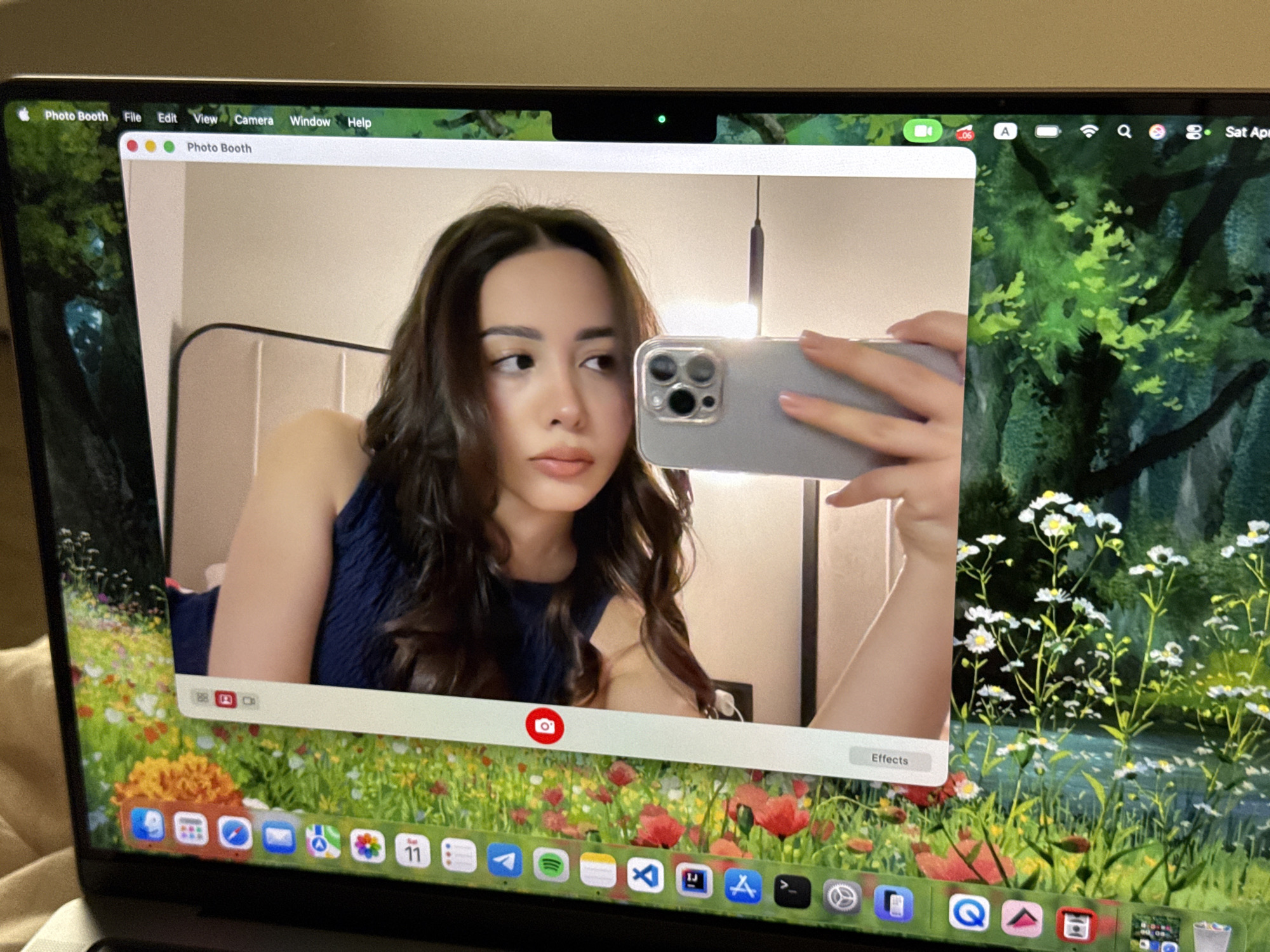 The image shows a laptop screen displaying the Photo Booth application. Within the application window, a young woman is taking a selfie with her smartphone. The background of the selfie appears to be a bedroom. The laptop's dock at the bottom of the screen shows various application icons. The laptop's background is a digitally enhanced nature scene. The laptop's top menu bar displays standard options like "File," "Edit," "View," "Camera," "Window," and "Help." The date "Sat Apr" is also visible on the top menu bar. There is a green light displayed above the application window, indicating that the laptop's camera is in use.
Location information is not present in the image.