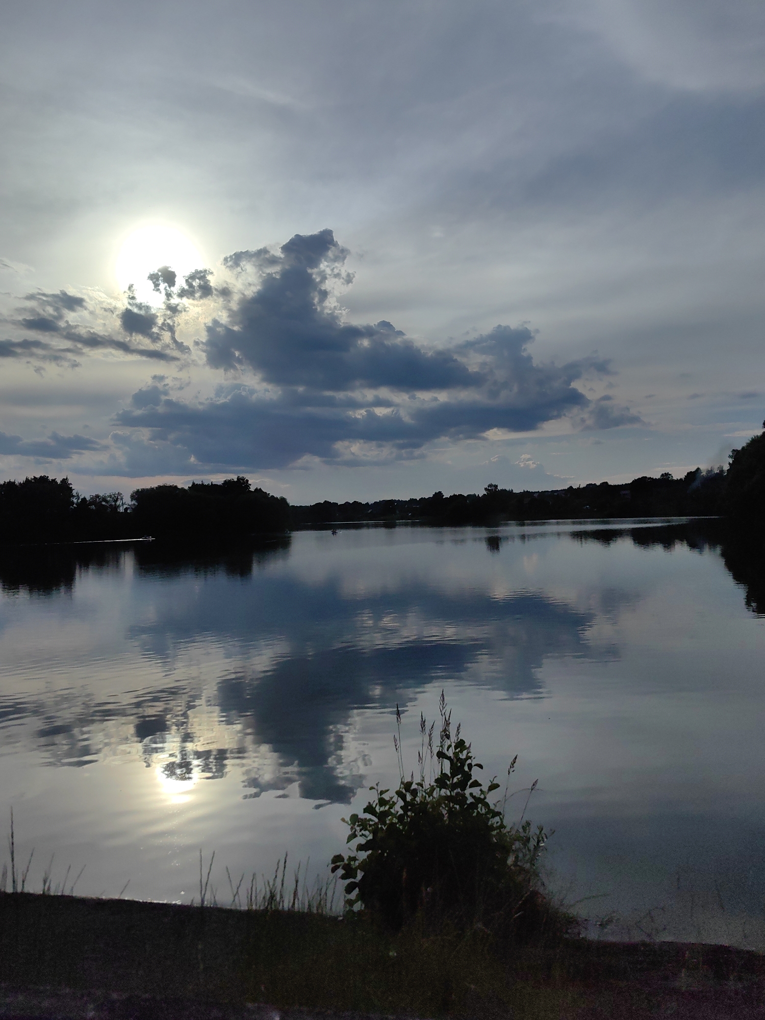 The image captures a serene lake scene at dusk, likely within the Дзямідавіцкі сельскі Савет, Belarus. The sun, partially obscured by dramatic, dark clouds, casts a shimmering reflection on the calm water. Silhouetted trees line the distant shore, forming a dark, undulating horizon. The foreground shows a patch of dark grass and foliage near the water's edge, with the water itself reflecting the sky and the sun's diffused light. No people or specific actions are depicted, creating a peaceful and tranquil atmosphere.