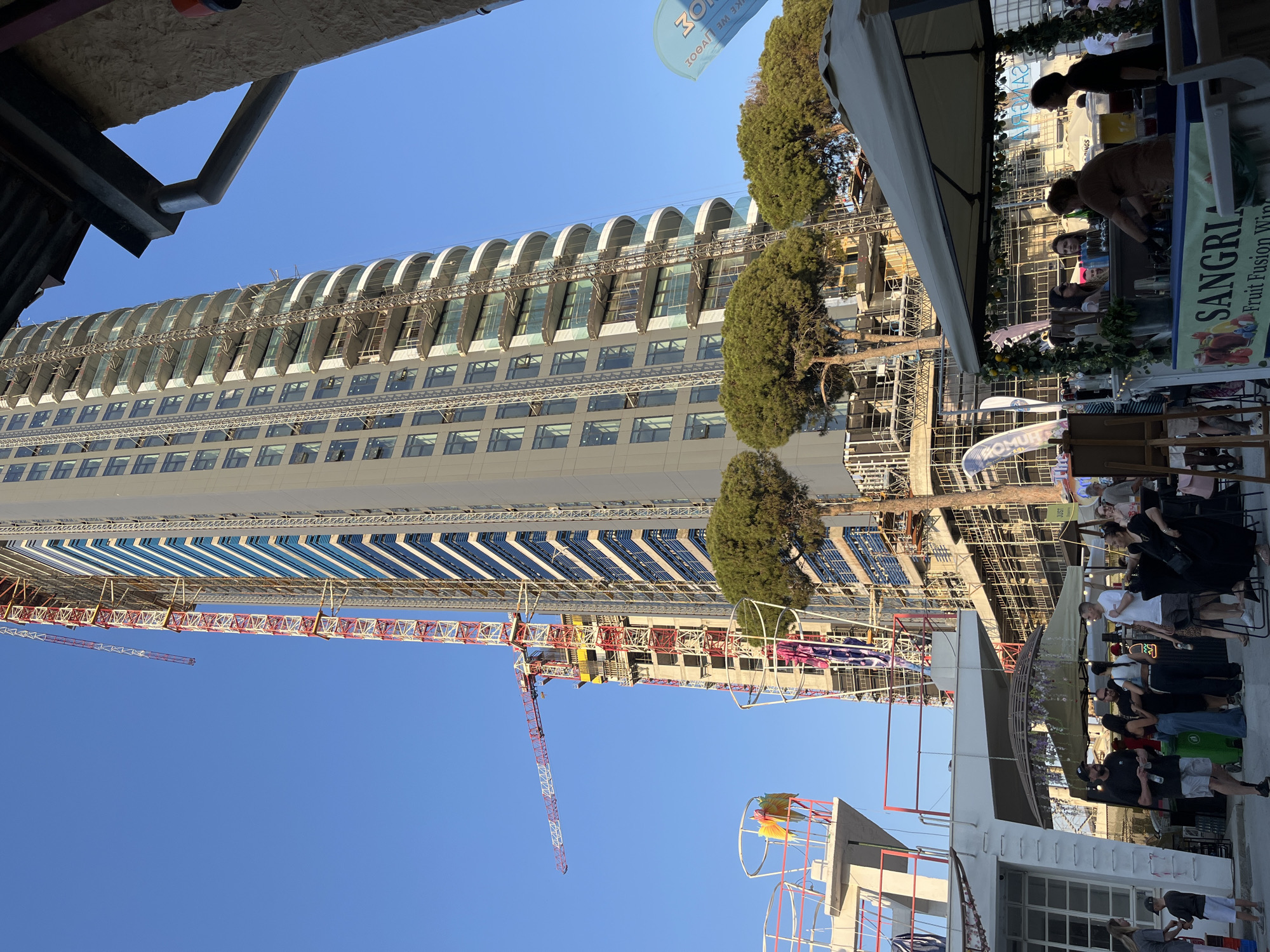 This detailed image depicts a bustling outdoor scene in the city of Limassol, Cyprus, under a clear blue sky on a bright, sunny day.
The dominant feature is a very tall, slender high-rise building under construction in the background. The building's facade on the right half is more completed, featuring numerous levels of curved, glass-fronted balconies and installed windows. The left half of the building reveals ongoing construction, with extensive scaffolding covering the structure and exposed concrete framework visible. Multiple red and white tower cranes are actively engaged in the construction, with one prominent crane extending its long jib horizontally on the left side, and another crane arm visible higher up.
In the midground and foreground, an outdoor market or social gathering is taking place. On the far right, a white canopy shades a stall displaying a "SANGRIA Fruit Fusion Wine" sign. Two individuals are positioned behind the counter, one appearing to serve drinks from large dispensers. A group of casually dressed people are gathered around this stall, some conversing. Further left, several other people are standing in groups, including adults and at least one child near a white, multi-level structure with red railings and a flat roof. This structure appears to be part of the foreground architecture.
Several short, manicured evergreen trees with dense, rounded canopies are situated between the market area and the construction site. A light blue banner or flag, possibly for an event or business, is partially visible on the far right and another further left. The ground appears to be a paved urban surface. In the upper right corner, a dark metal gutter and a section of a stone pillar from an overhead structure are visible. The overall scene suggests a vibrant urban environment combining new development with public leisure activities.