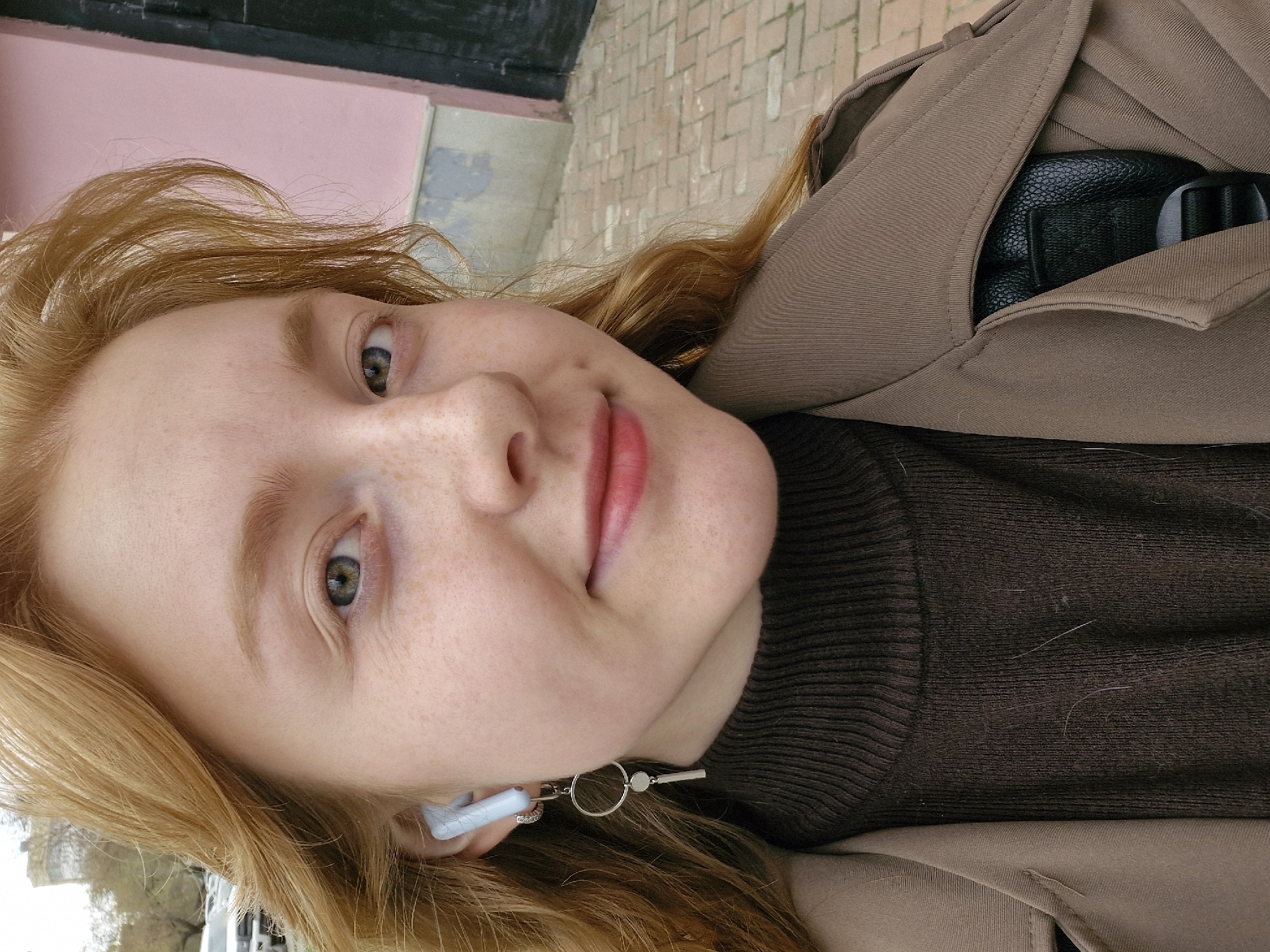 A medium close-up, portrait-style photograph captures a young woman with long, wavy, reddish-blonde hair, fair skin, and numerous freckles. Her head is tilted to the left, and she gazes directly at the camera with a gentle, friendly smile. Her eyes are a striking green or hazel, and she is wearing light pink lipstick.
She is dressed in a dark brown ribbed turtleneck sweater layered beneath a light brown or tan trench coat. Distinctive details include light-colored earbuds in her left ear, from which a decorative earring, featuring a small ring and a key-like charm, dangles. Partially visible within the pocket of her trench coat is a black, textured object, likely a camera or a compact bag.
The background indicates an outdoor setting during the daytime, with soft, diffused lighting. A section of a light pink wall or building facade is visible behind her head. To her right, there is a textured surface resembling light-colored brickwork or paving stones. The image conveys a calm and approachable mood. There is no visible text in the photograph. This image was taken in Yekaterinburg, Russia.