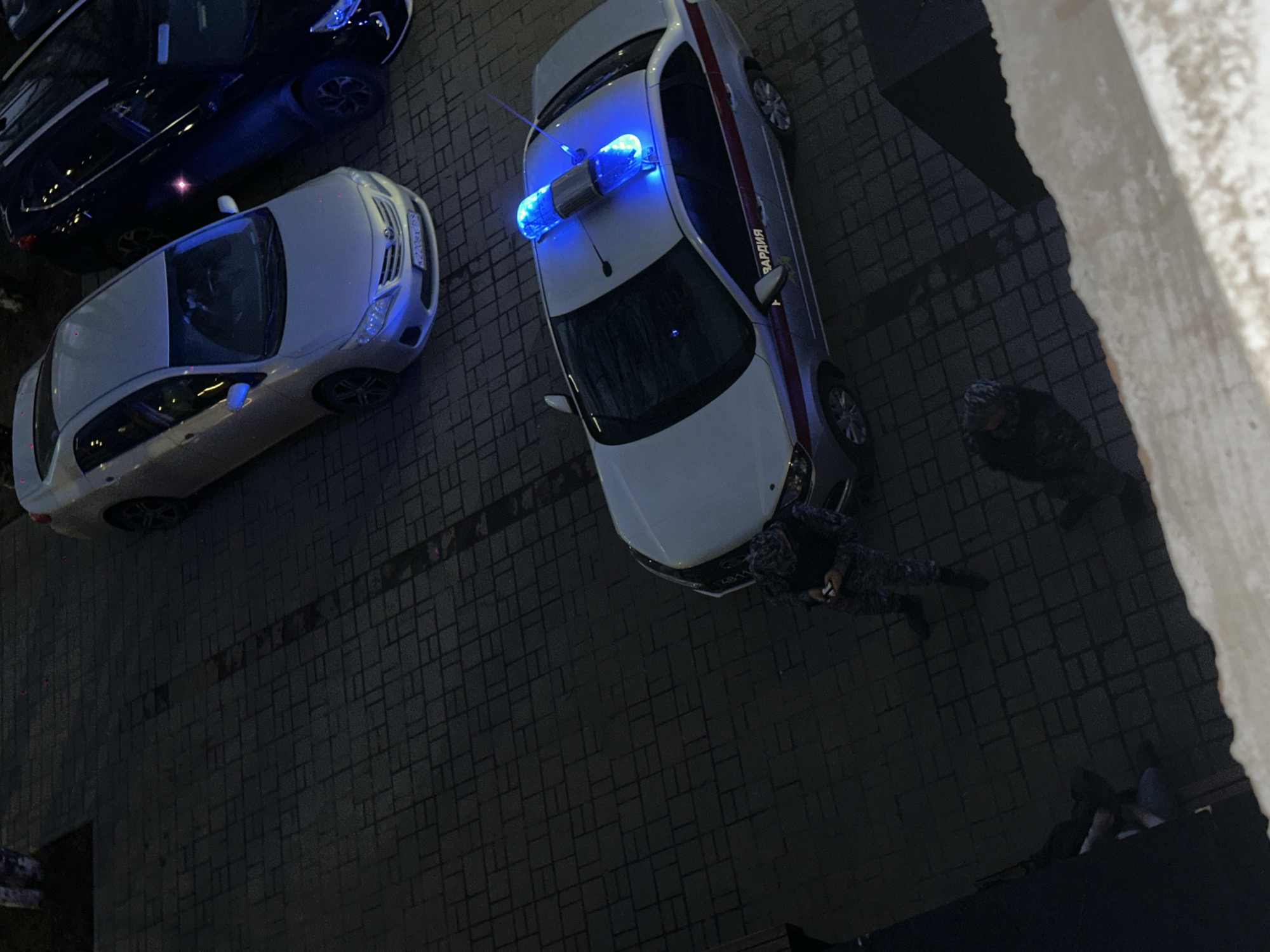The image shows an aerial view of a street in Glazov, Russia, with three parked cars. One is a police car with blue lights flashing. Two individuals, possibly law enforcement officers, are near the police car. The environment consists of a brick-paved surface, with part of a building visible on the right side of the frame.