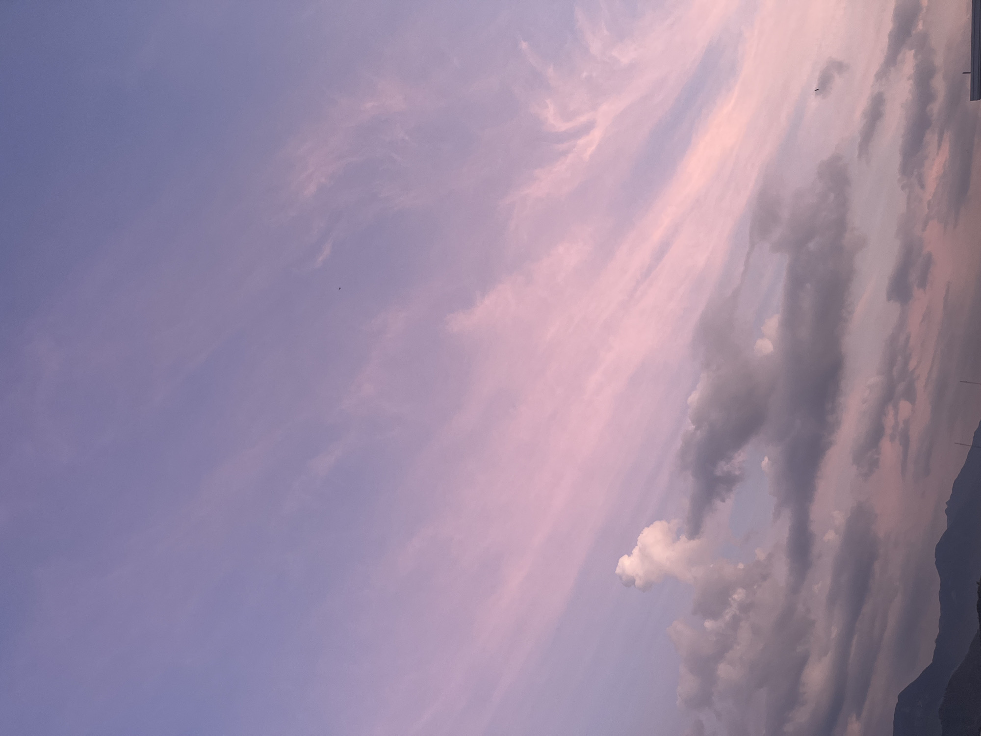 The image shows a sky view in what is possibly the city of Bac Nha Trang Ward, Vietnam, during twilight or sunrise. The sky is predominantly a light purple hue, interspersed with streaks of pink and white cirrus clouds. Cumulus clouds are present towards the bottom right corner, near a dark mountain range silhouette. A few distant, unidentified flying objects can be detected. The image is rotated.